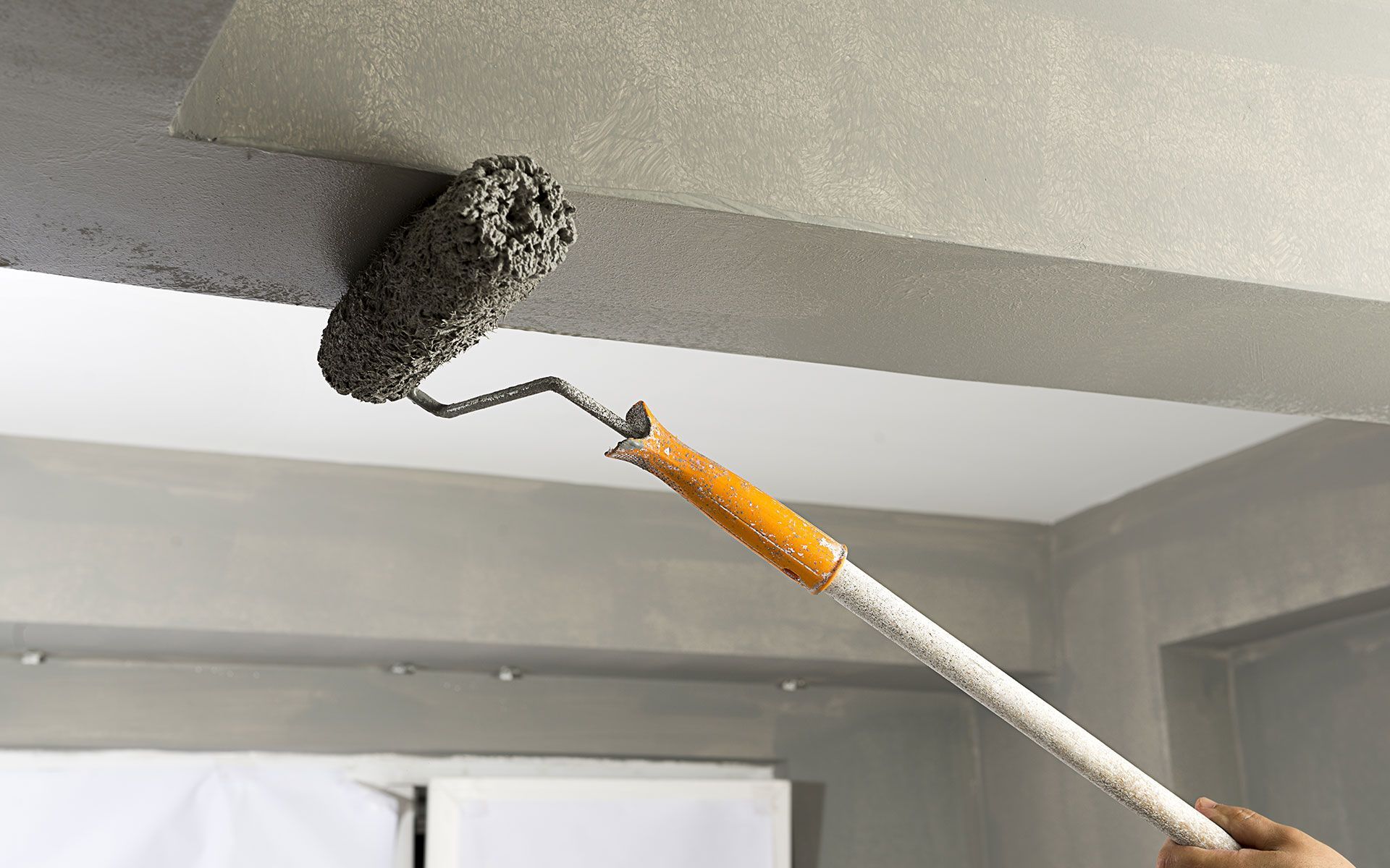 Person painting a ceiling with a roller, holding a long handle. Grey paint on roller, indoor setting.