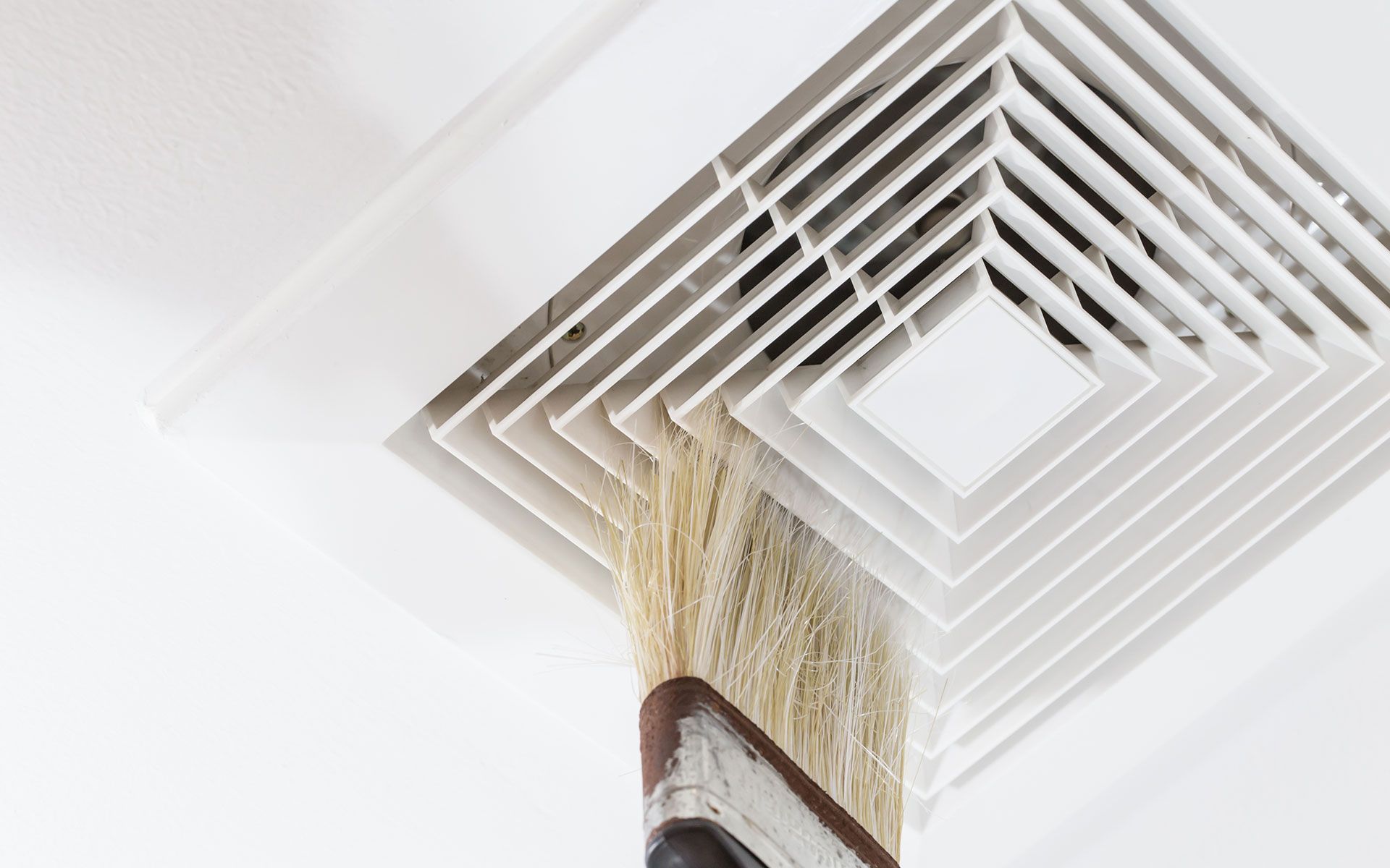 A person is cleaning a dusty ceiling vent with a brush. White ceiling and vent, brown and gray brush.