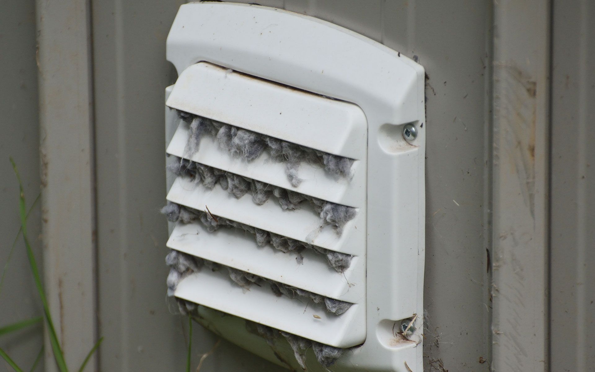 White dryer vent cover clogged with lint on a gray wall.