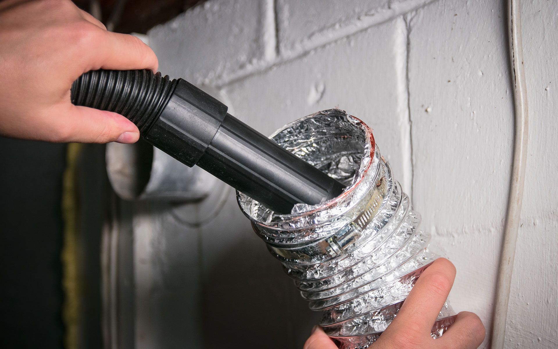Hands vacuuming out a dryer vent. Black hose inserted into silver, flexible ducting on a white wall.