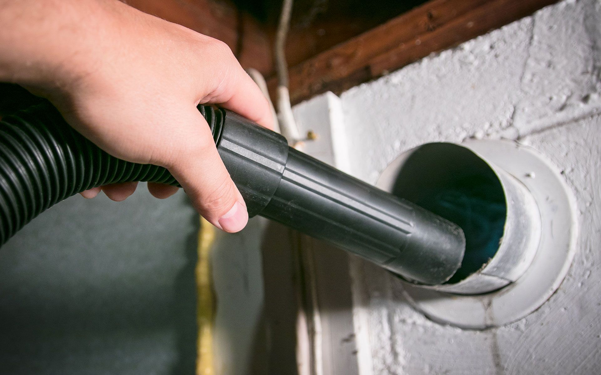 Hand holding vacuum nozzle cleaning a white air duct opening in a wall.