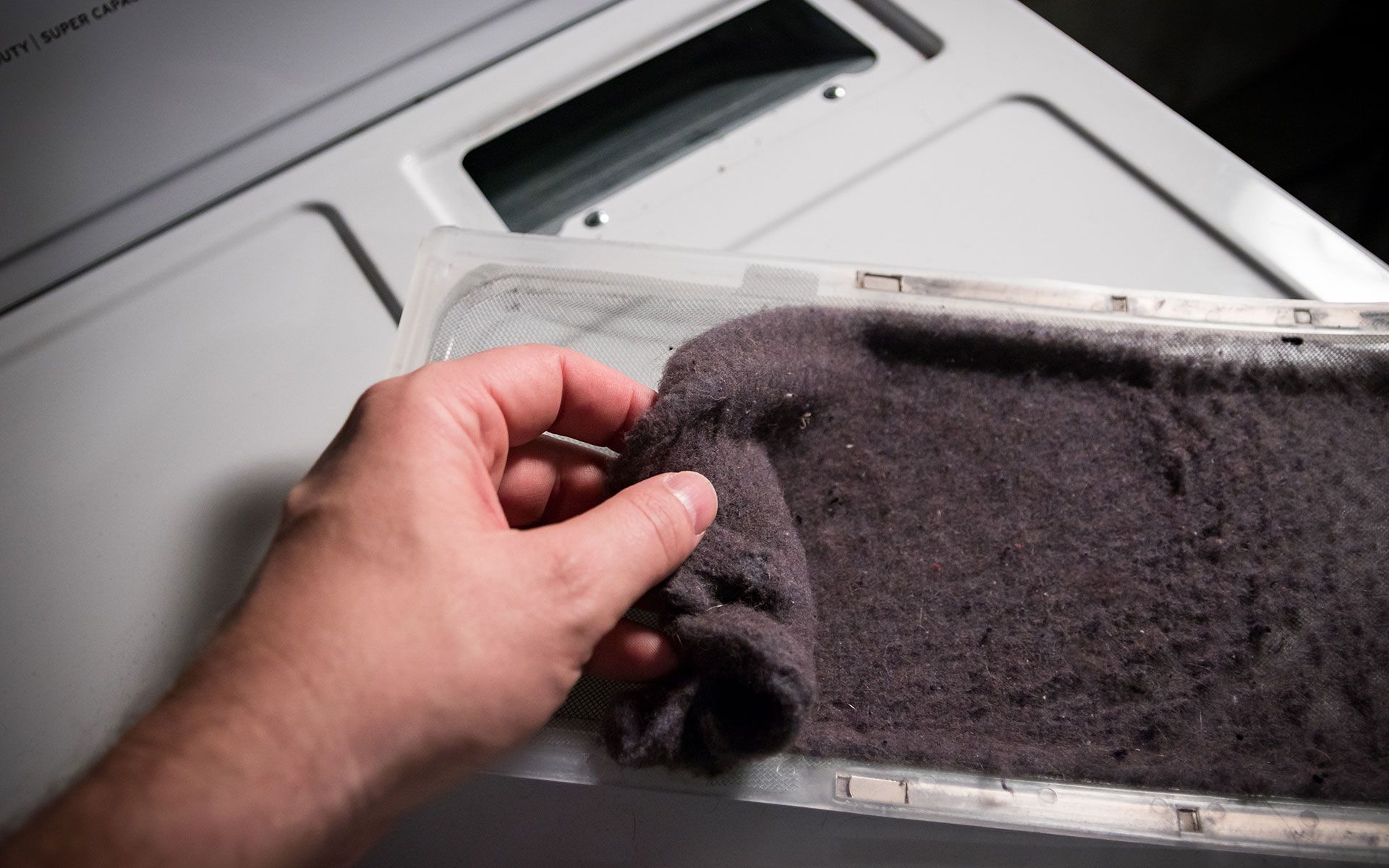 Hand holding a thick, dark gray lint screen pulled from a white dryer.
