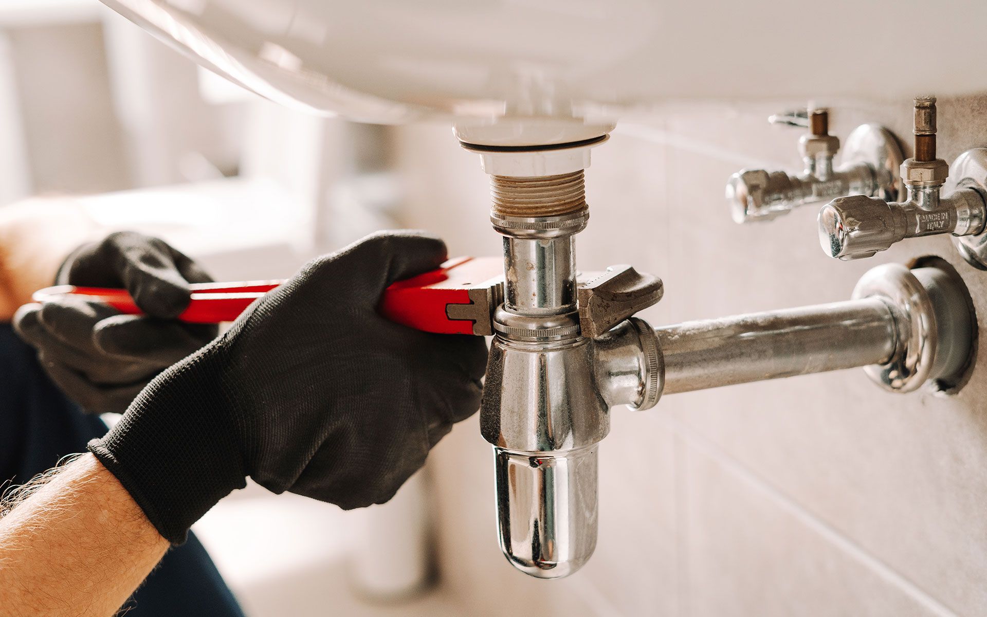 Plumber using a wrench to work on pipes under a white sink, wearing black gloves.