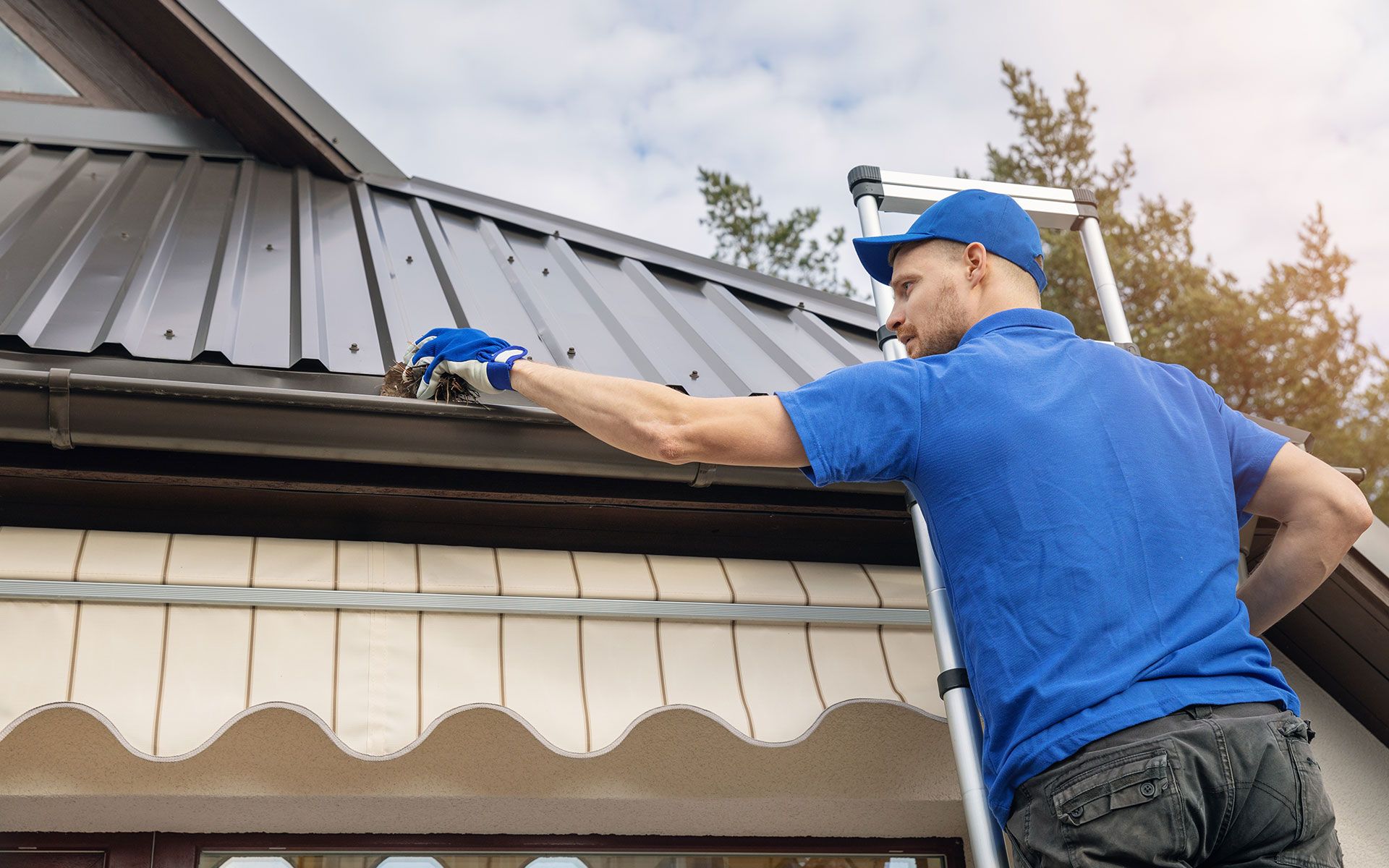 Man on ladder cleaning gutters on a house with dark roof and white trim.