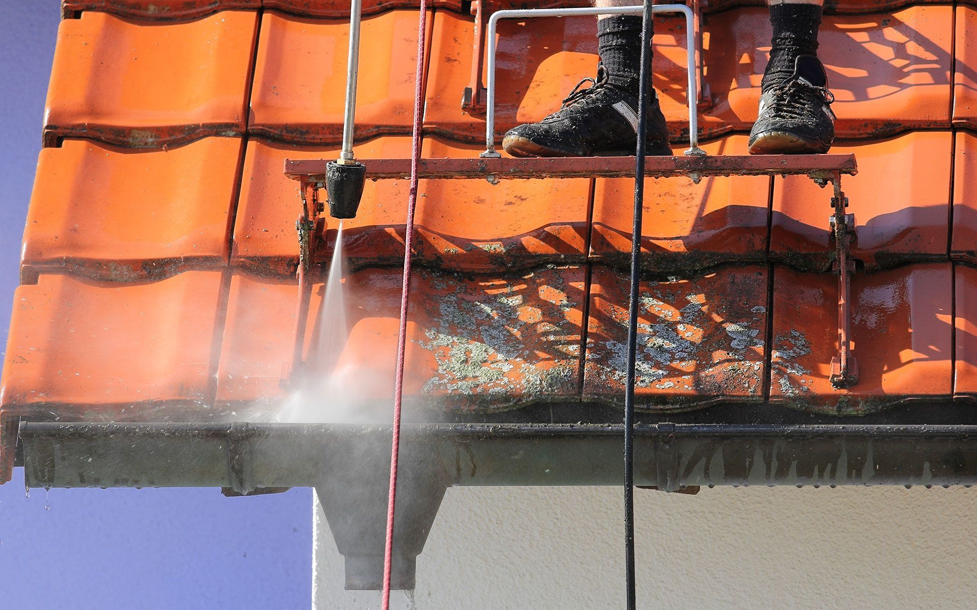 Person on ladder pressure washing roof's gutter. Water sprays, cleaning debris. Red tiles, blue sky.