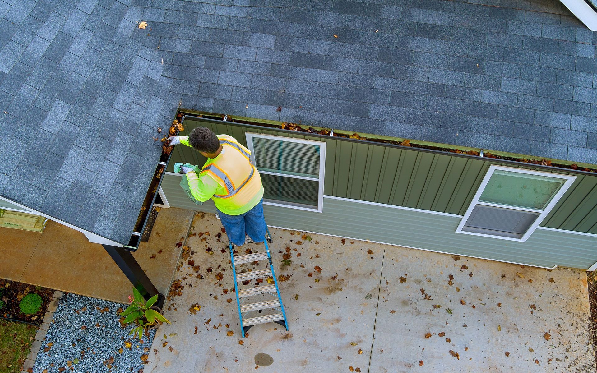 Person on ladder cleaning leaves from a roof gutter of a house.