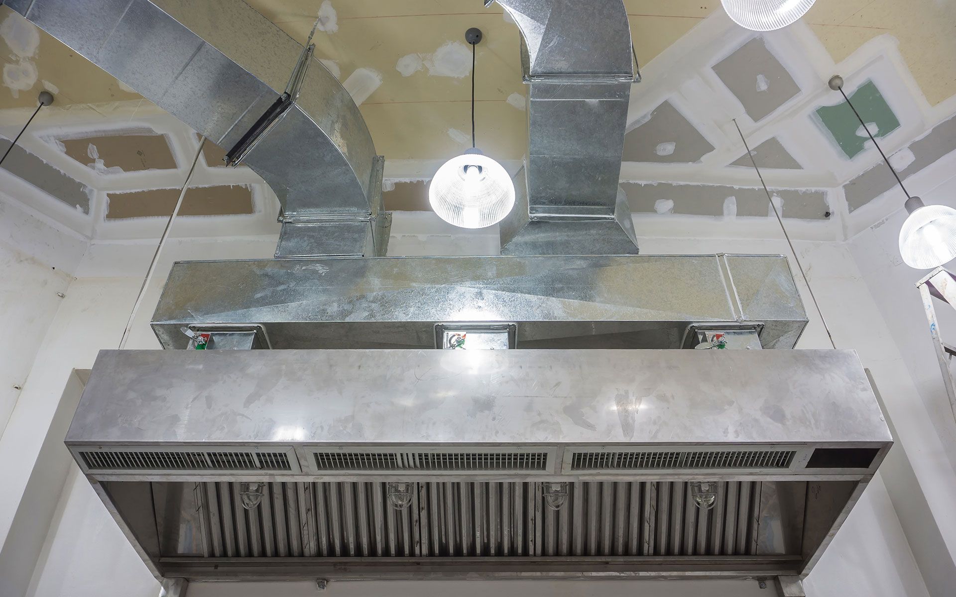 Stainless steel commercial kitchen exhaust hood with ductwork, suspended from a textured ceiling.