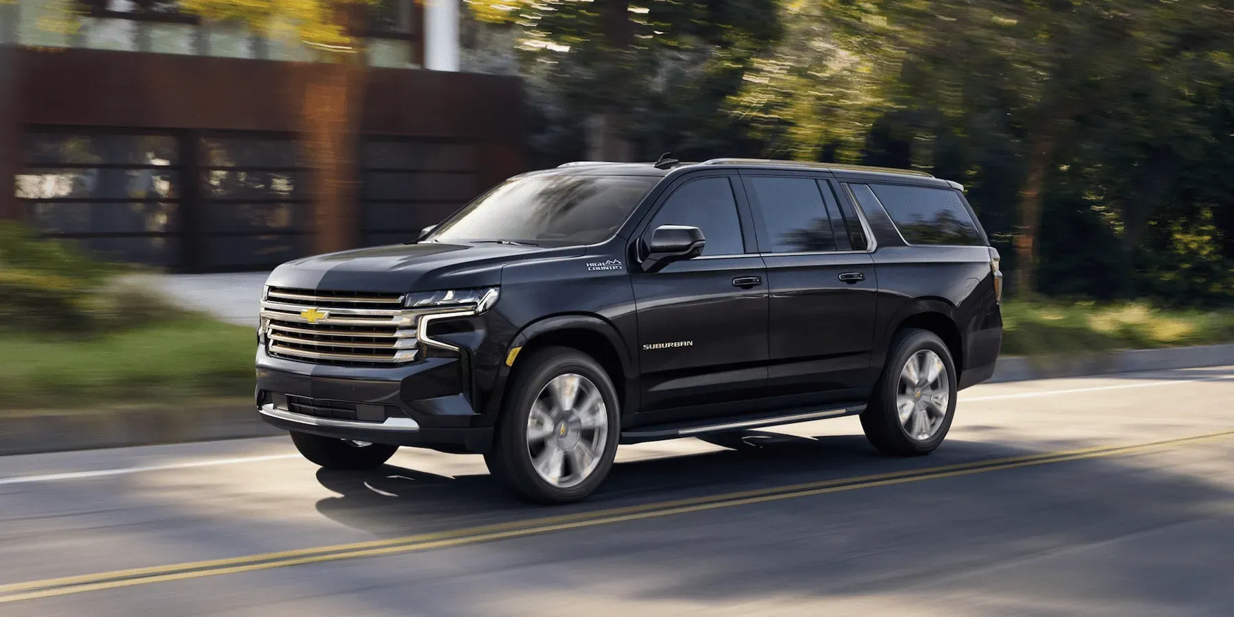 Black Chevrolet Suburban SUV on a paved road, driving near a building and trees.