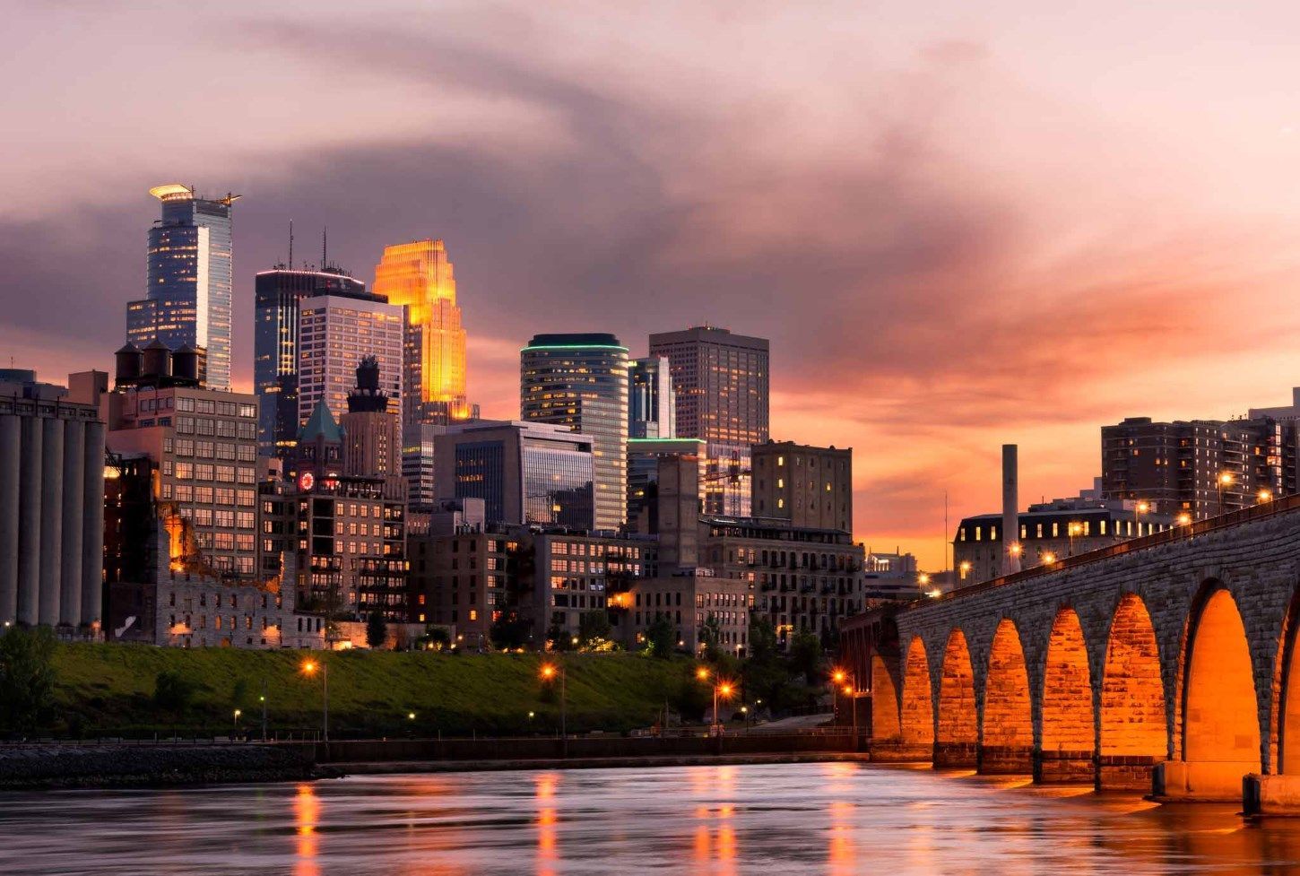 Skyline of Minneapolis at sunset, buildings and stone arch bridge reflecting in the river.