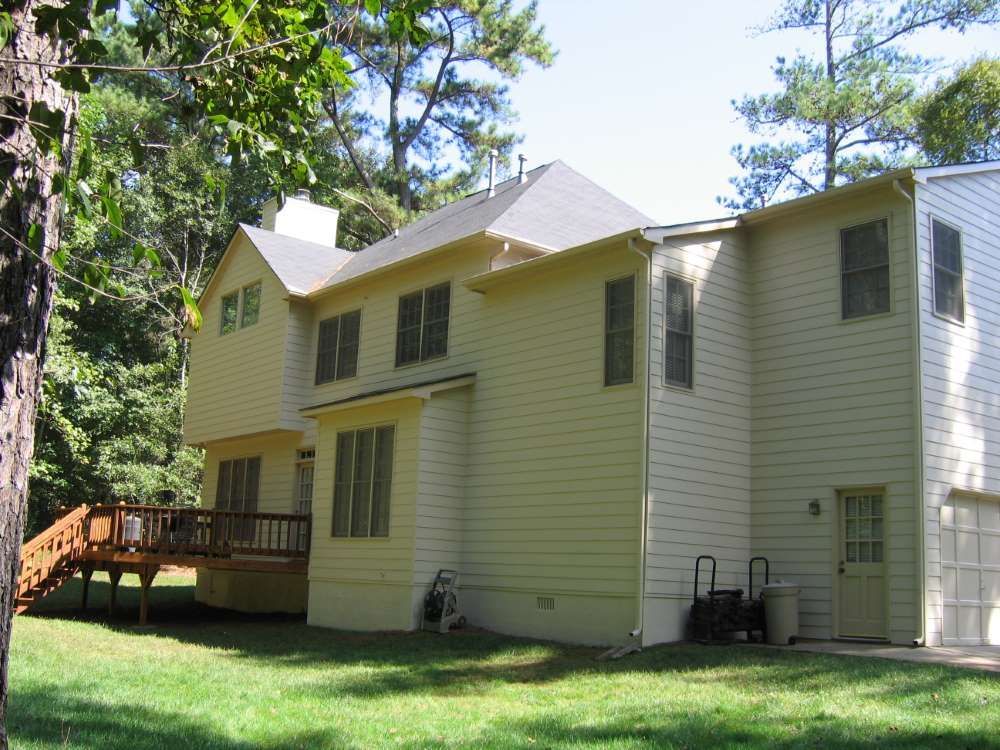 A large yellow house with a deck and trees in the background