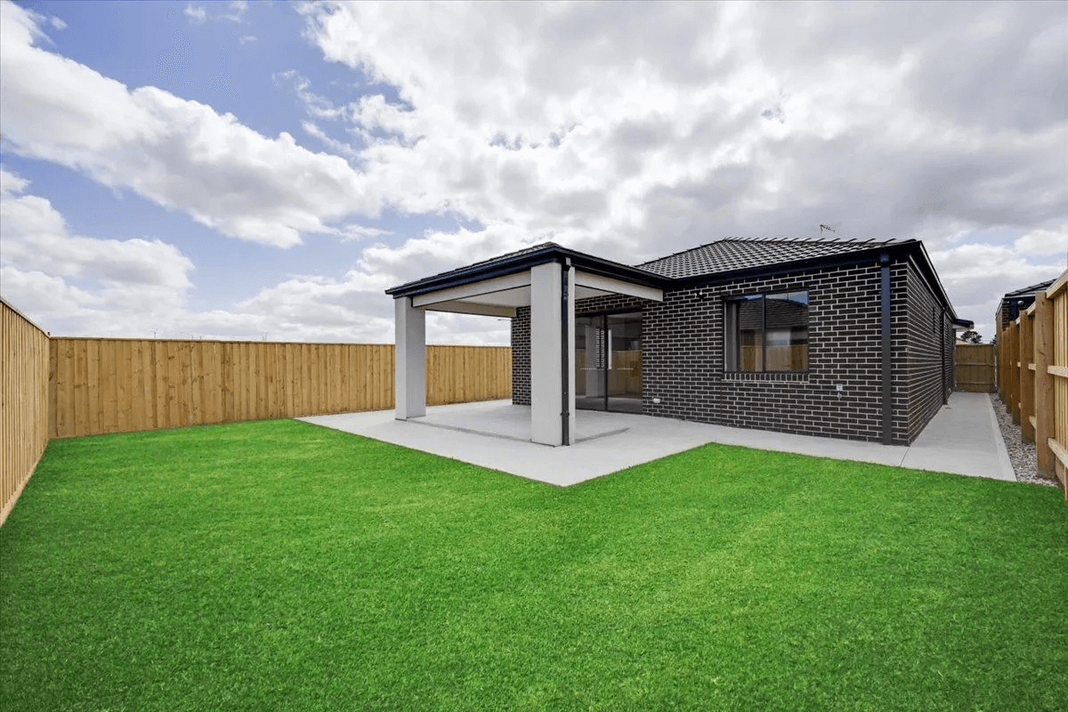 A modern brick house with large windows and a covered patio sits in a fenced backyard with bright green grass on Denmead Drive, Donnybrook, VIC 3064, under a partly cloudy sky.