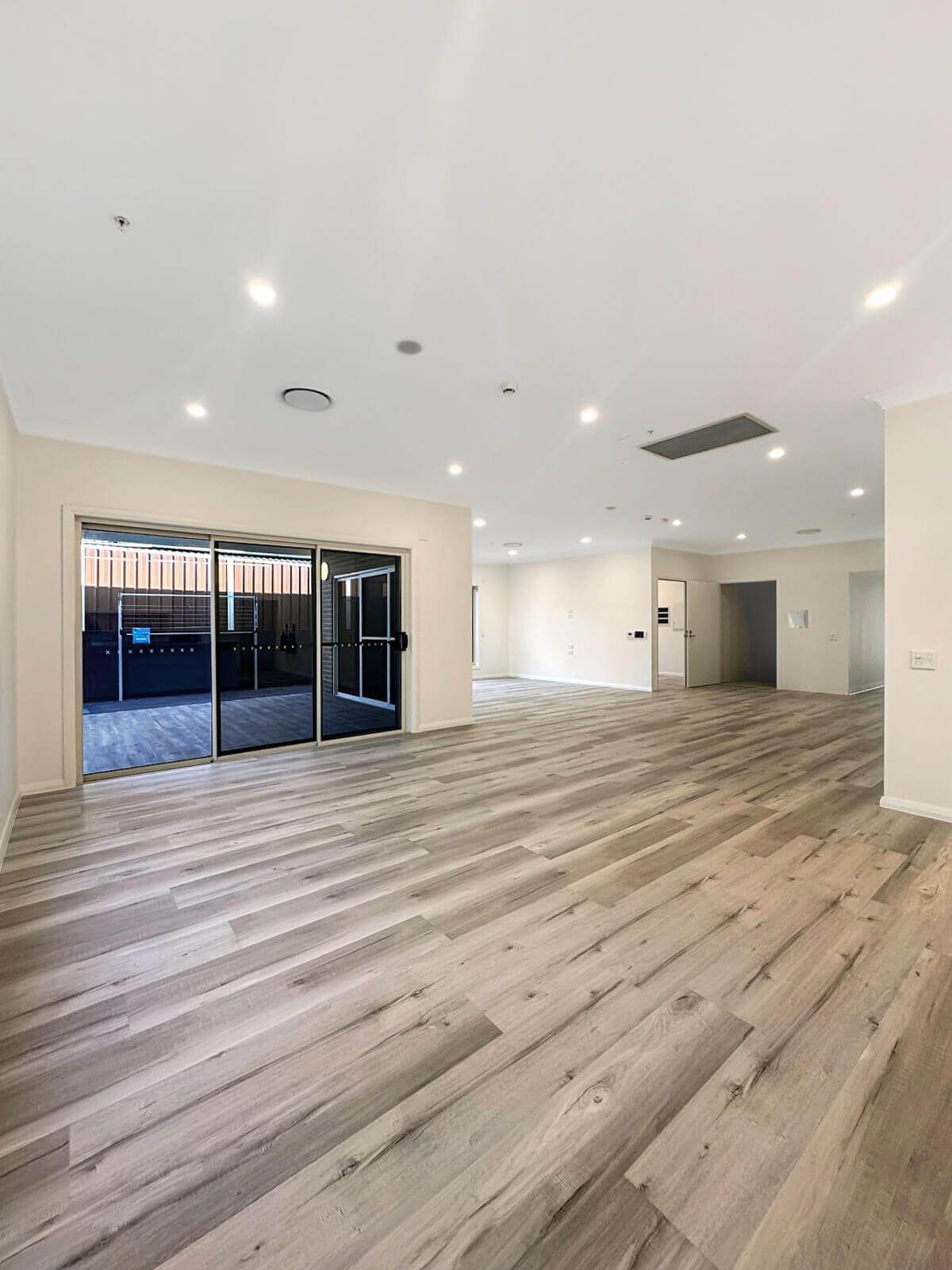 Spacious, empty room with light wood flooring and white walls on Hamlet Crescent in Rosemeadow NSW. Recessed ceiling lights add ambiance, while large glass sliding doors lead to a fenced patio. The room seamlessly extends towards another unfurnished space in the background at 2560.