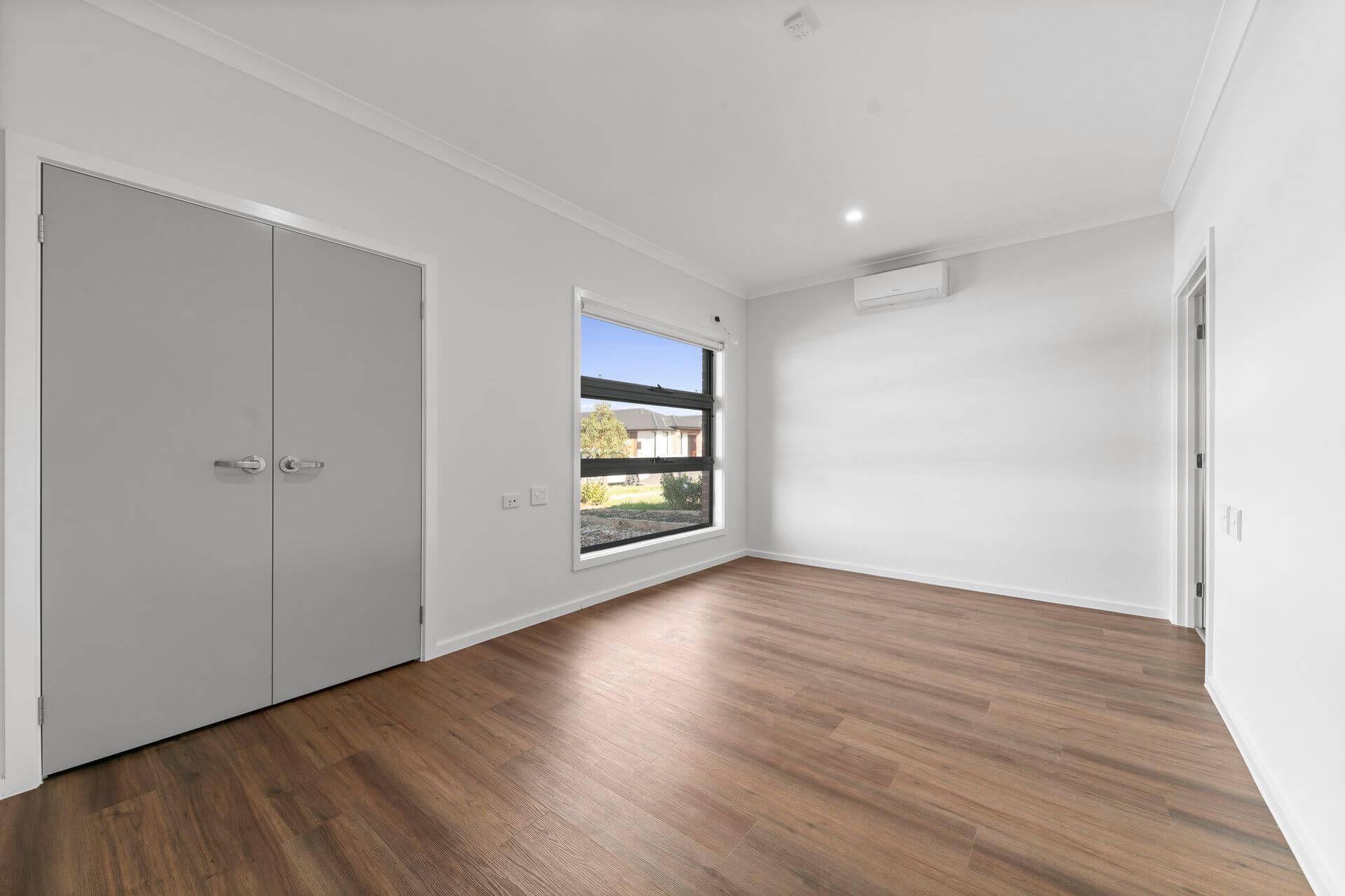 Minimalist, empty room with white walls and wood flooring in Bonnie Brook, VIC 3335. A large window fills the space with natural light. Features double gray closet doors, overhead ceiling light, and a wall-mounted air conditioning unit on Canyon Circuit.