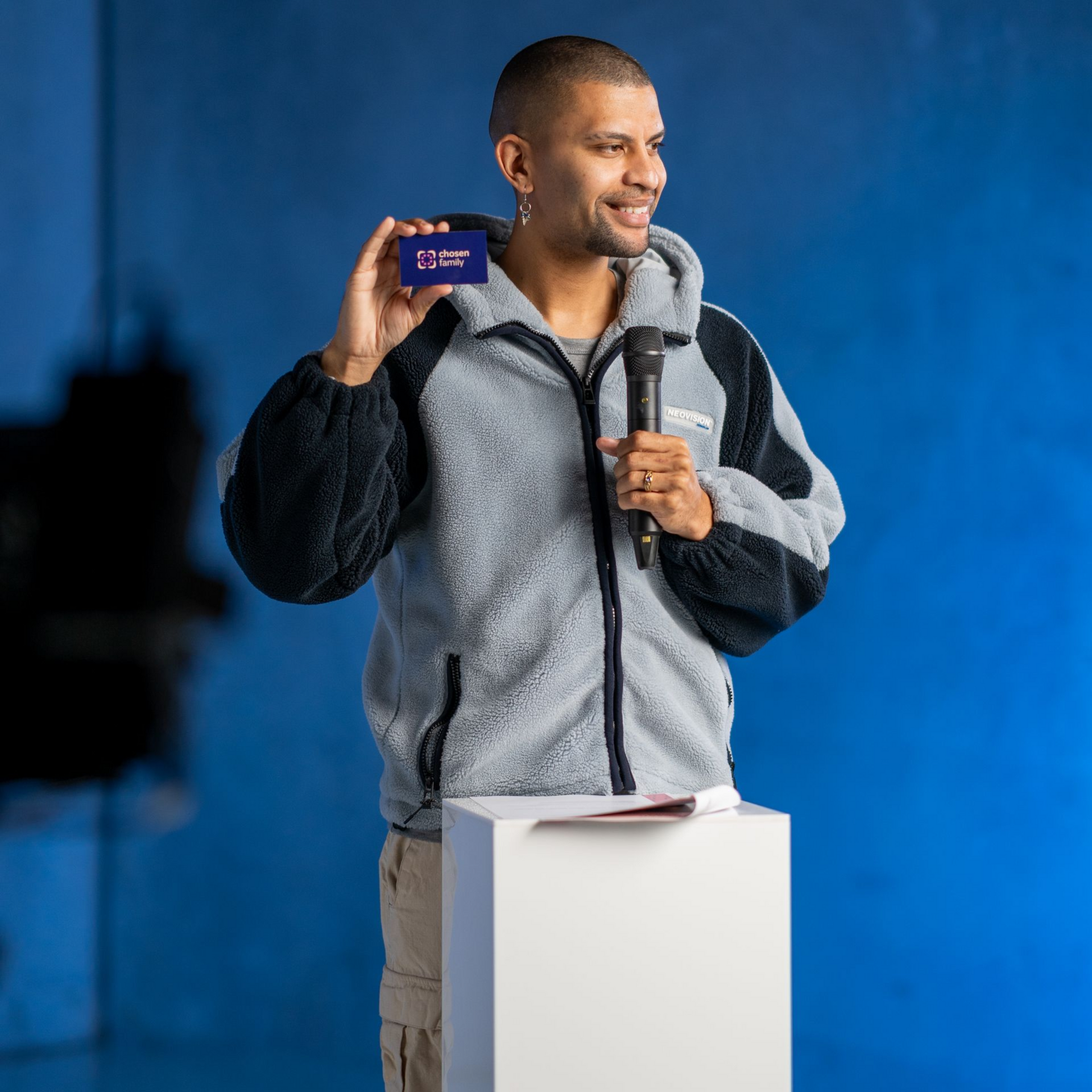 Man holding card, speaking into microphone. He stands behind white pedestal against a blue backdrop.
