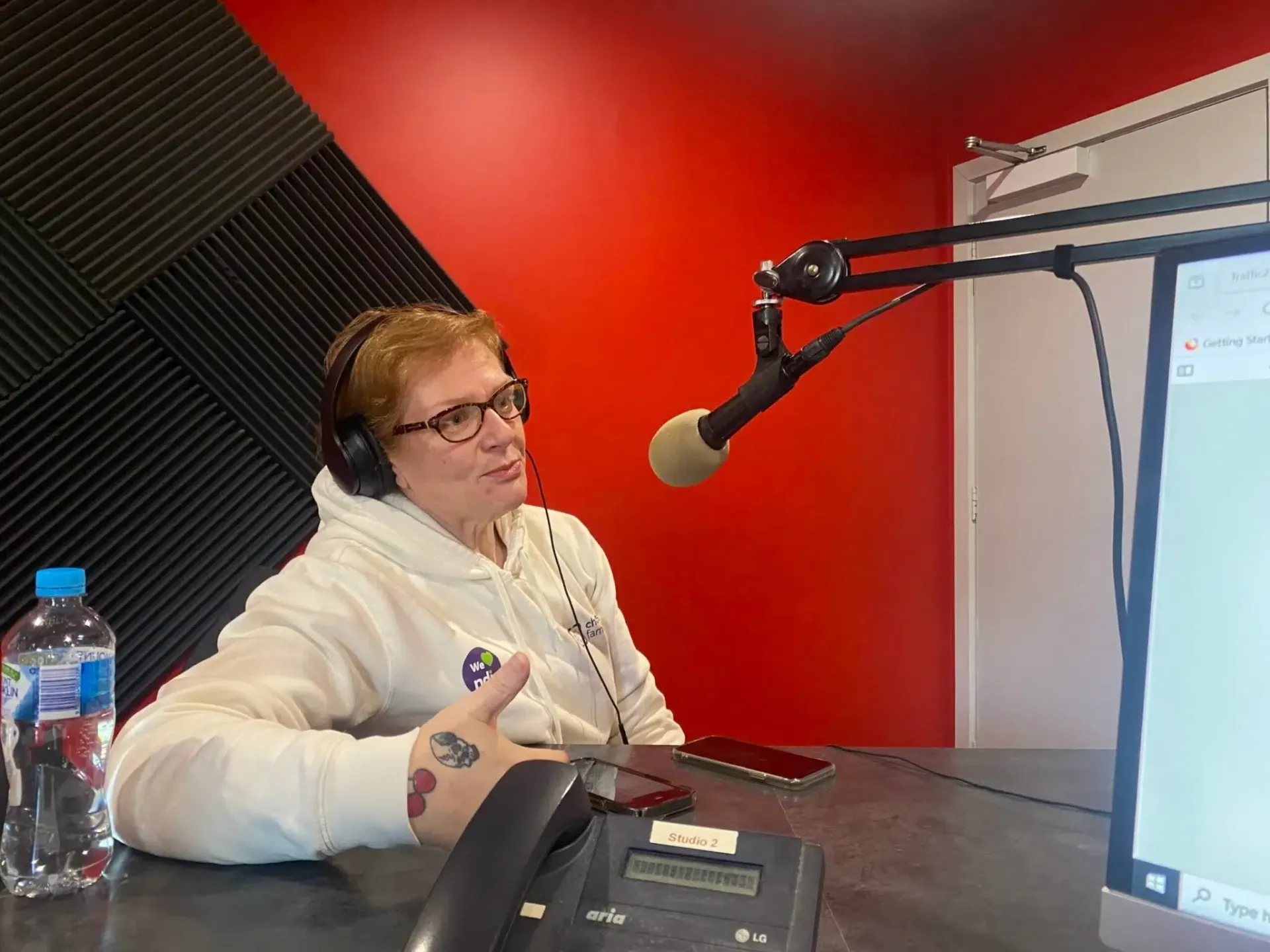 Person wearing glasses and headphones speaks into a microphone in a red-walled studio.
