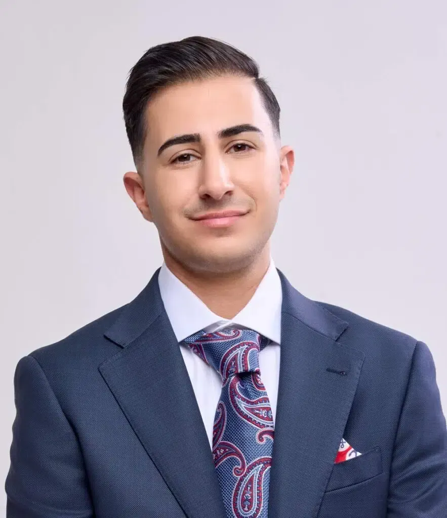 Man in a suit smiles, tie with paisley pattern, small red pocket square, light background.