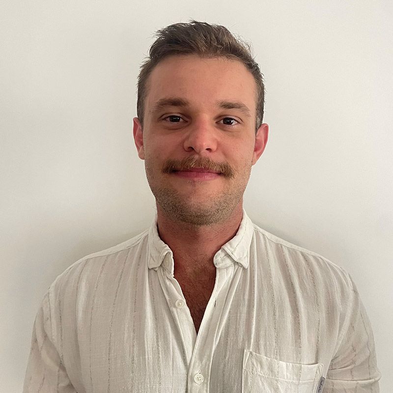 Man with a mustache wearing a light-colored button-up shirt smiling. White wall background.