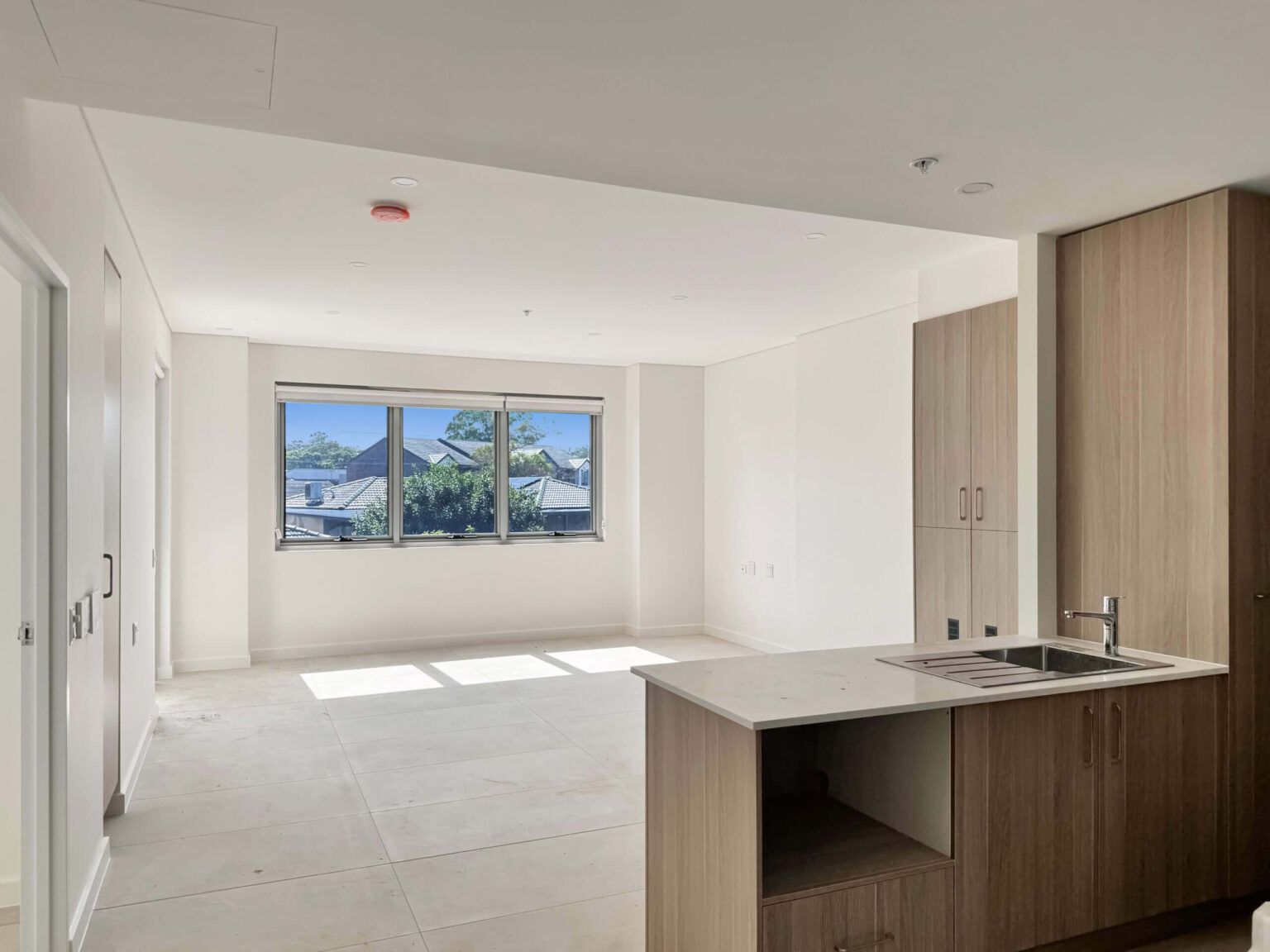 The remaining modern apartment interior at Leumeah Apartments features white walls and tiled floors. An open-plan kitchen with wooden cabinets stands to the right, while large windows ahead allow natural light to fill this perfect space for single occupancy.