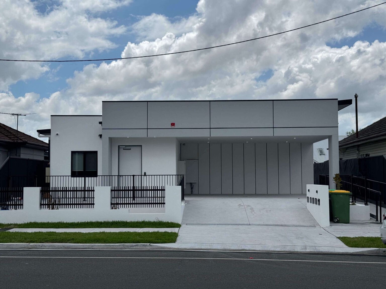 A modern white villa on Louis Street boasts a flat roof and clean lines, complemented by a covered driveway and sleek black railing. The partly cloudy sky frames this Granville gem, with a green bin by the entrance and neighboring houses on either side.