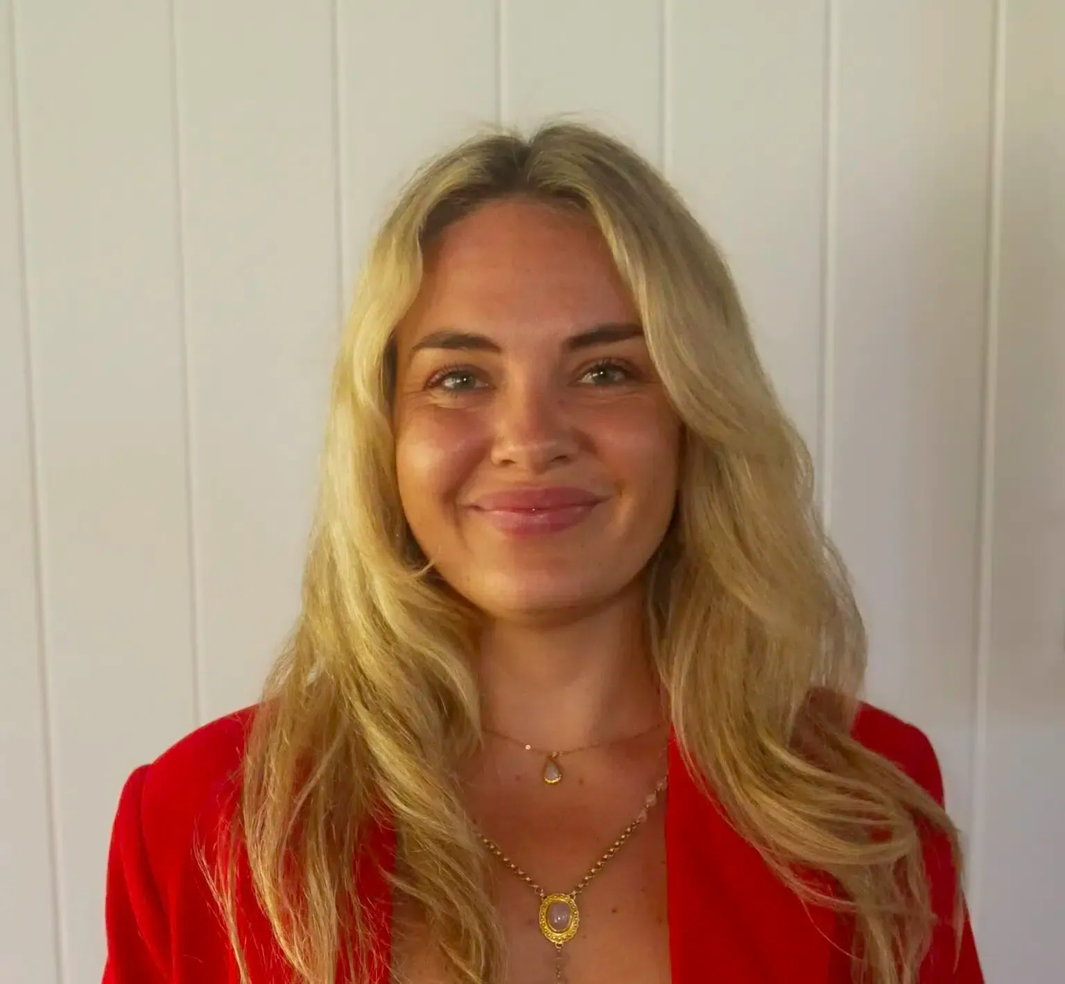 Woman with blonde hair wearing a red blazer smiling at the camera, against a white wall.