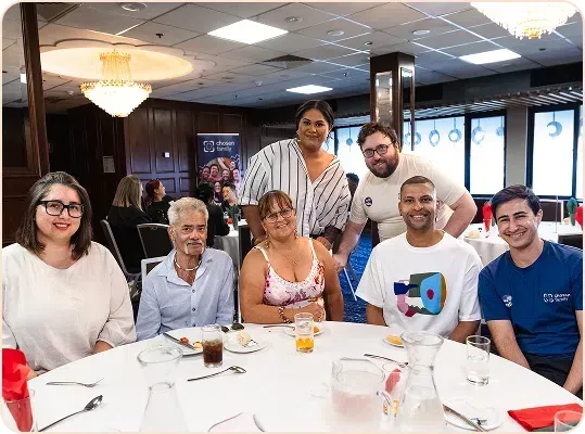 Group of people seated at a round table, smiling. Restaurant setting with food and drinks visible.
