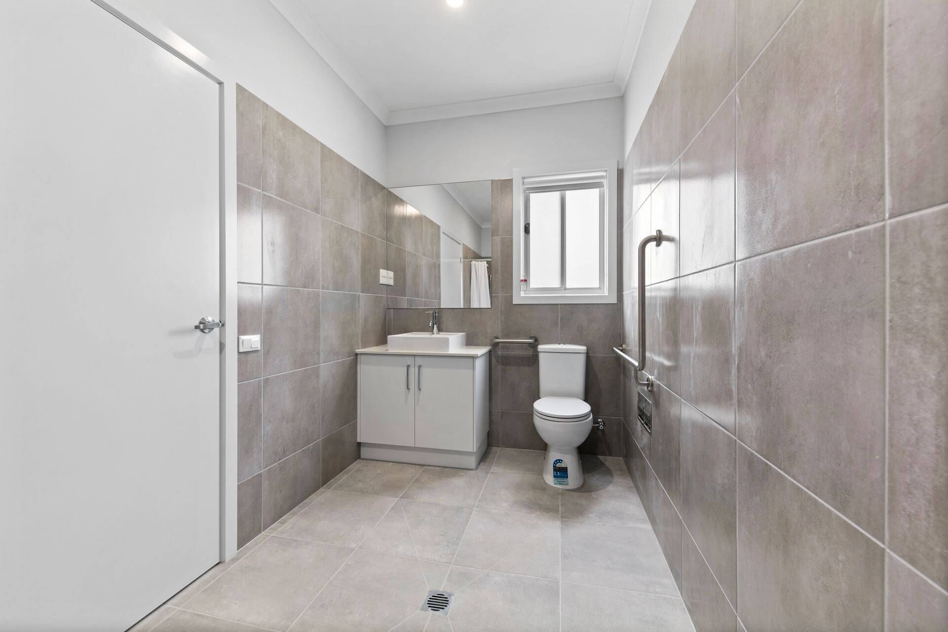 Modern, spacious bathroom with beige tiles, a white sink and cabinet, wall-mounted mirror, toilet, grab bar, and a window. Located in Weir Views, VIC 3338 near Edgewater Boulevard. A cleaning product sits on the floor next to the toilet.