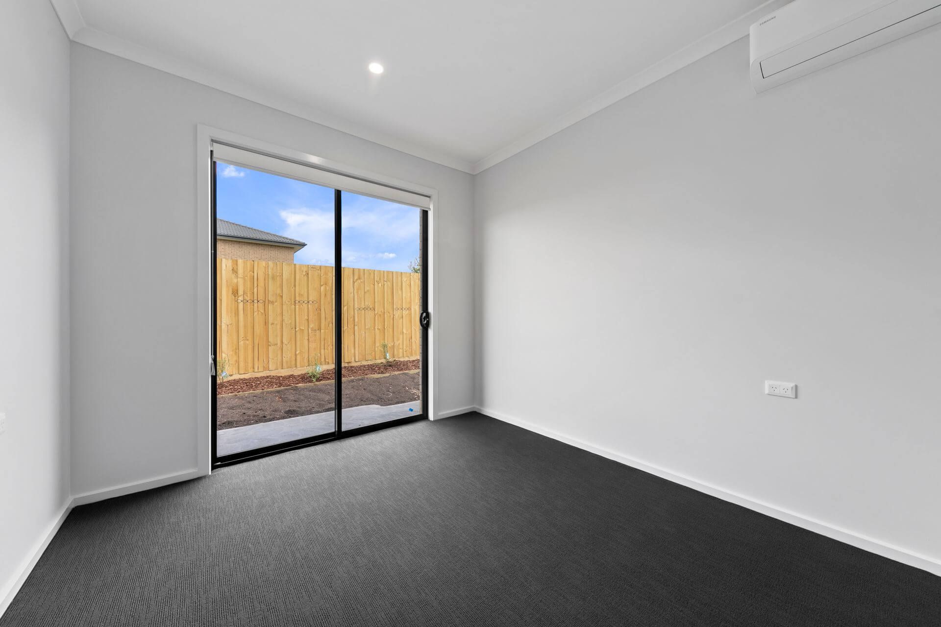 A bright, empty room with grey carpet, white walls, a sliding glass door to a fenced yard, and wall-mounted air conditioner in Weir Views, VIC 3338. Located near Marriott Boulevard for convenient access to local amenities.