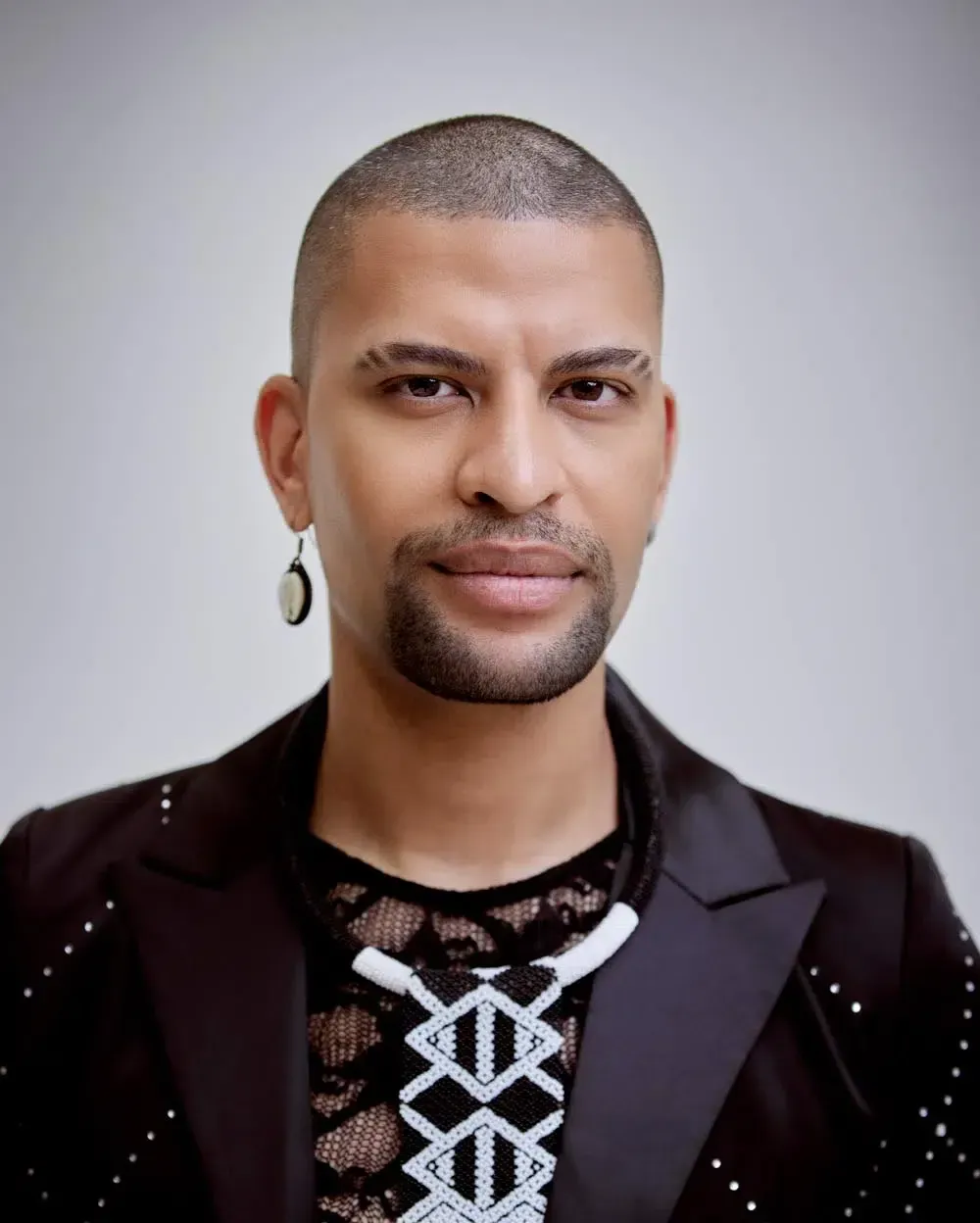 Man with shaved head, earring, and black blazer with studded detail.