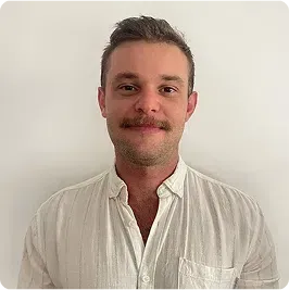 Man with a mustache, wearing a white button-down shirt, smiles at the camera against a white background.