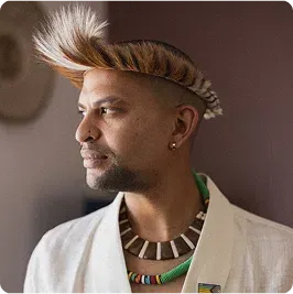 Man wearing traditional Zulu headgear, beaded necklaces, white jacket.