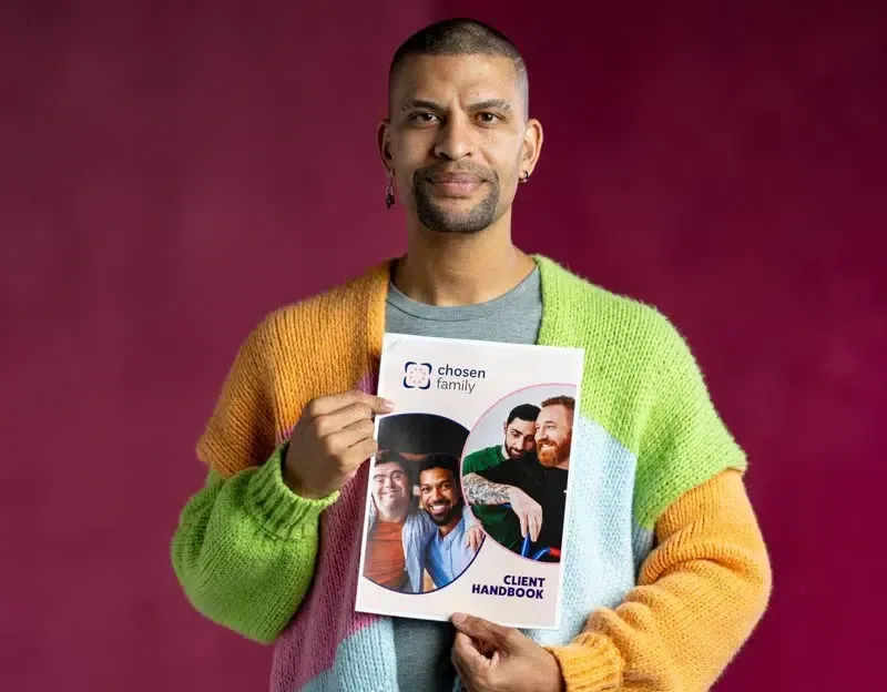 Man holding a document, wearing colorful sweater, smiles. Document features photos of three men.