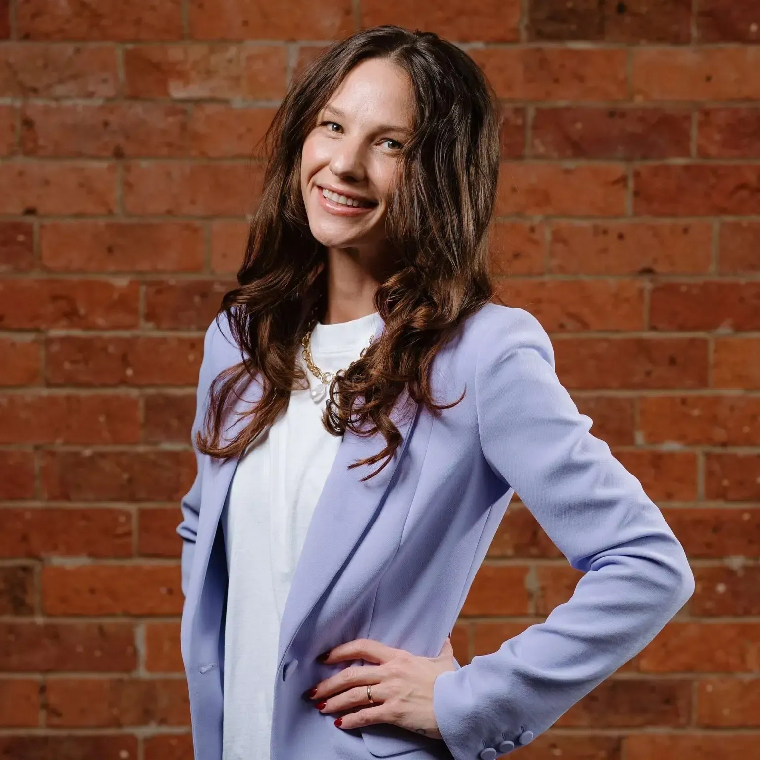 Woman in light purple blazer smiles, hand on hip, in front of a brick wall.