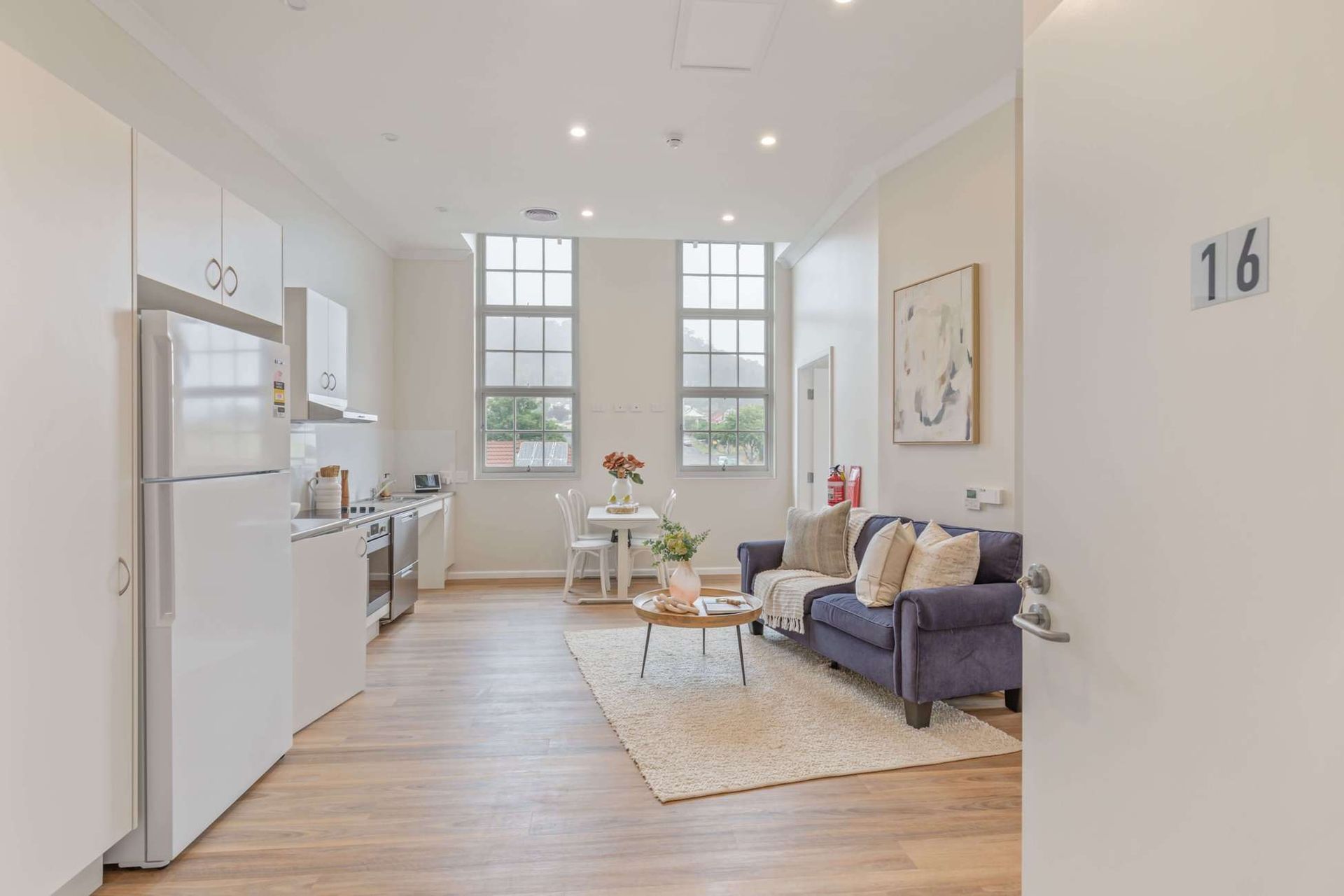 Experience the charm of Lithgow Apartments, where modern design meets luxury. This bright space boasts high ceilings, a kitchen with sleek white cabinets, a floral-adorned dining table, and a cozy blue sofa on a beige rug. Natural light pours through expansive windows for an inviting atmosphere.