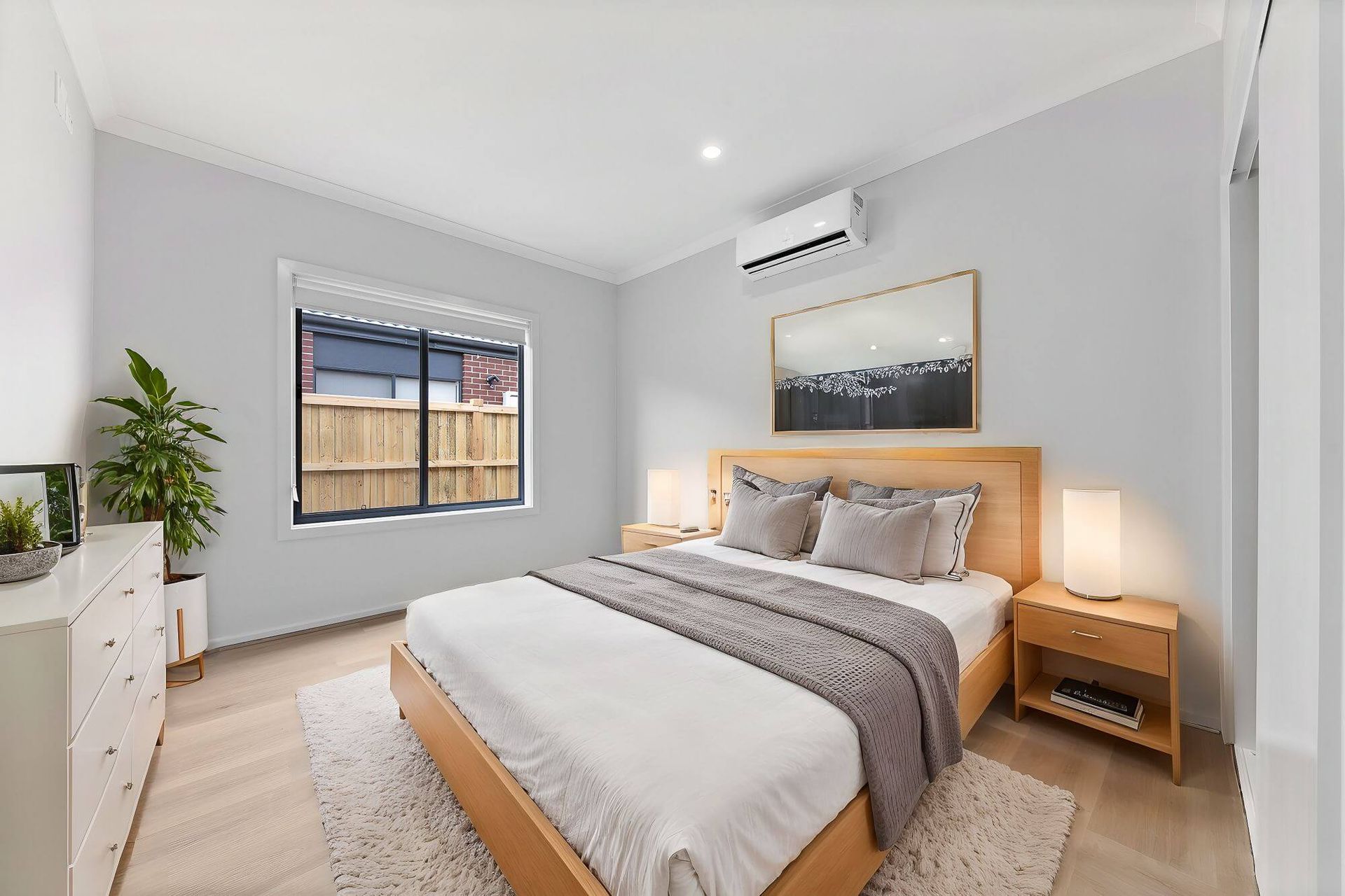 The modern bedroom, part of the Mandalay Estate collection, features a large bed with gray and white bedding on a wooden frame. Two bedside tables with lamps flank the bed. A potted plant and dresser enhance the left side, while a window with blinds and wall-mounted air conditioning complete this elegant space.