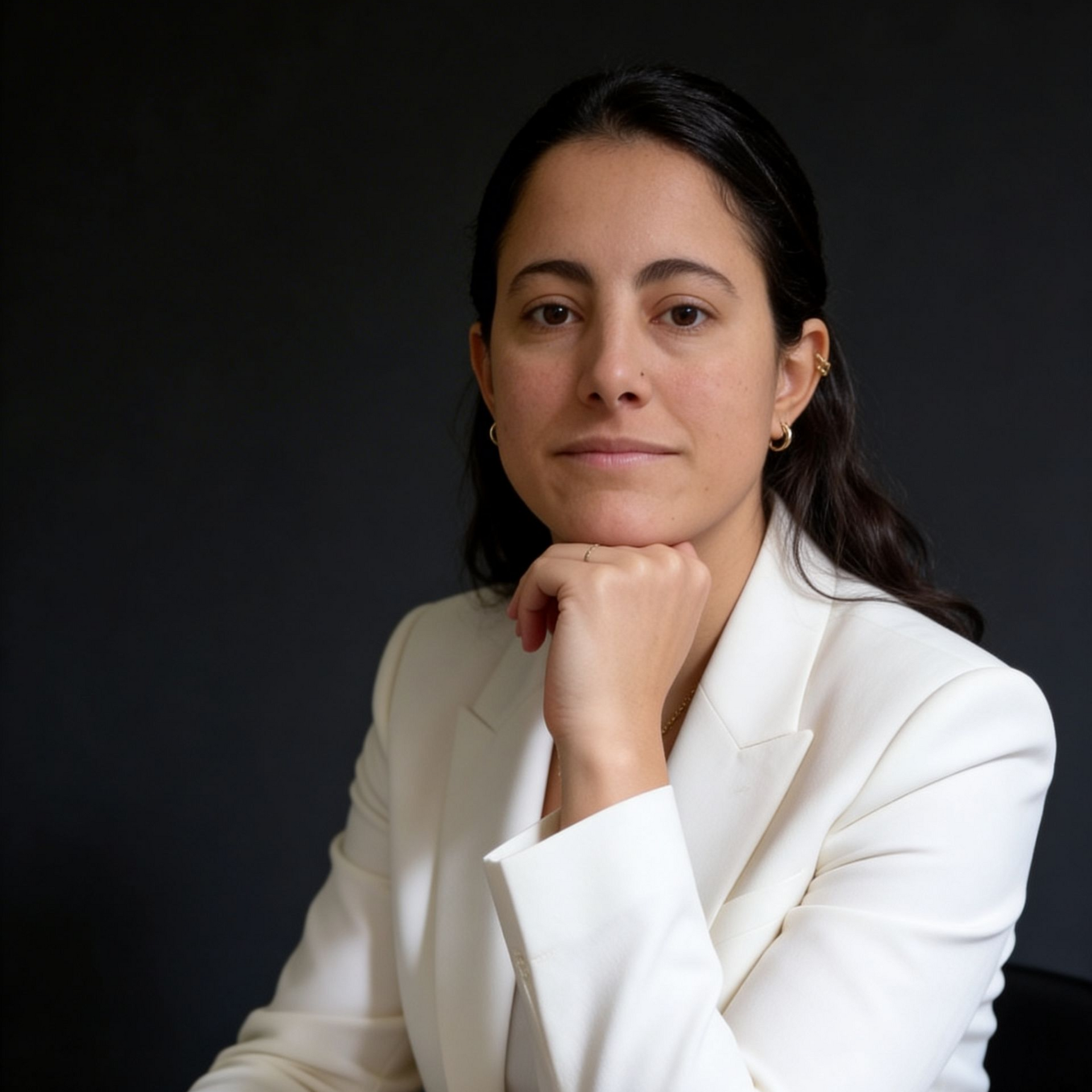 A person wearing a white blazer sits at a dark table with their chin resting on their hand against a black background.