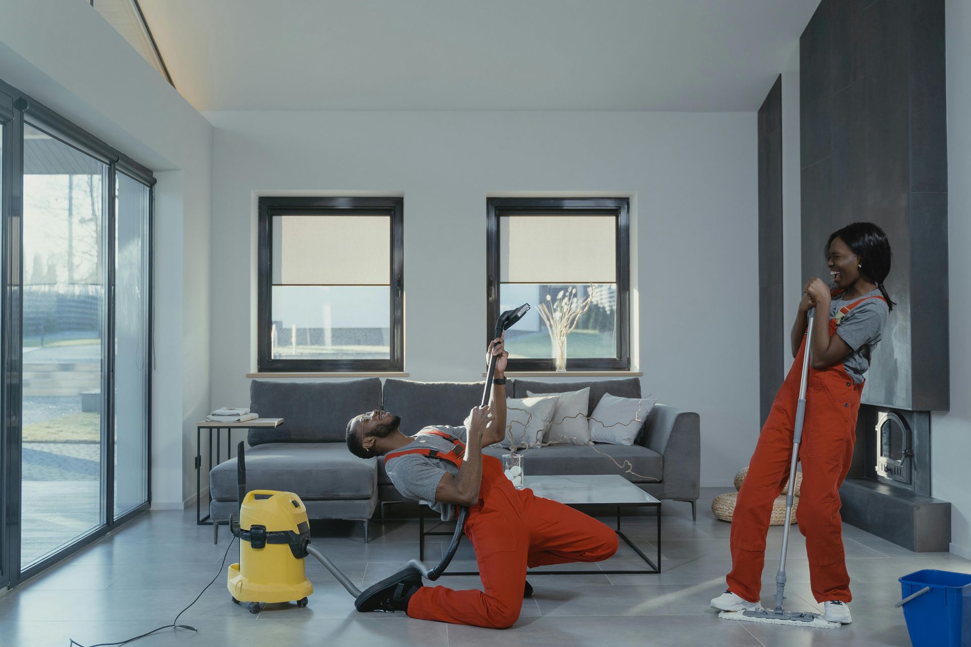Two people cleaning a modern home playfully. One pretends a vacuum is a guitar. The other sings into a mop.