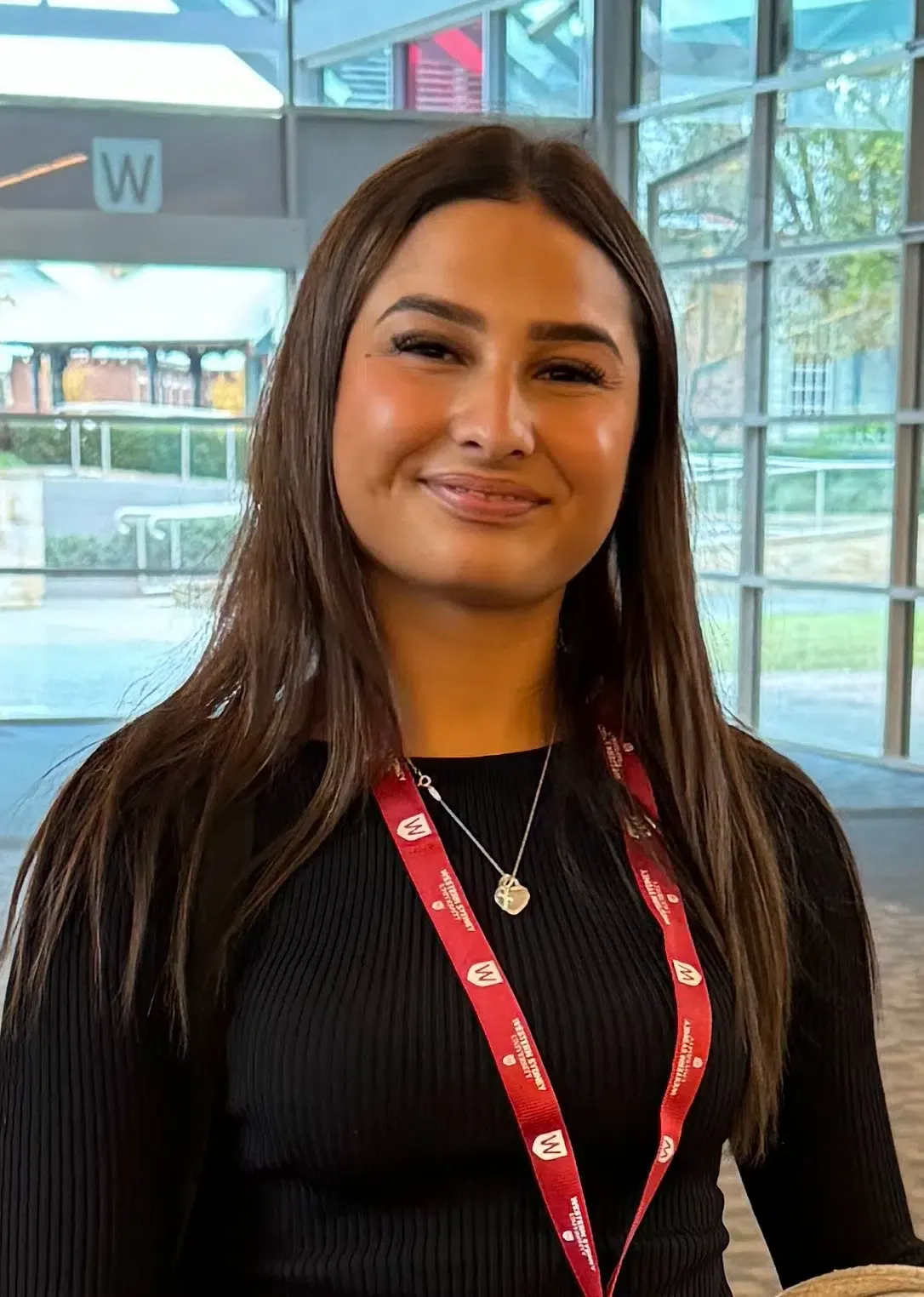 Woman with long brown hair wearing a black top and a lanyard, smiling indoors.