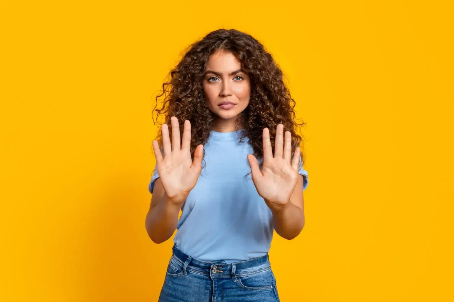Woman with curly hair holds up both hands, palms out, in front of a yellow background.