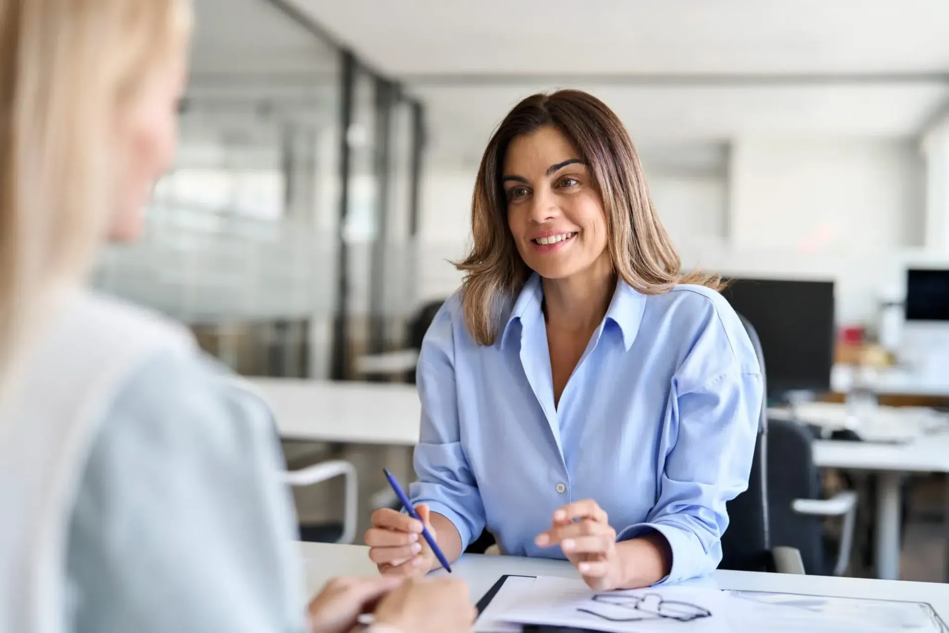 Woman in a blue shirt smiles, interviewing someone at an office desk.