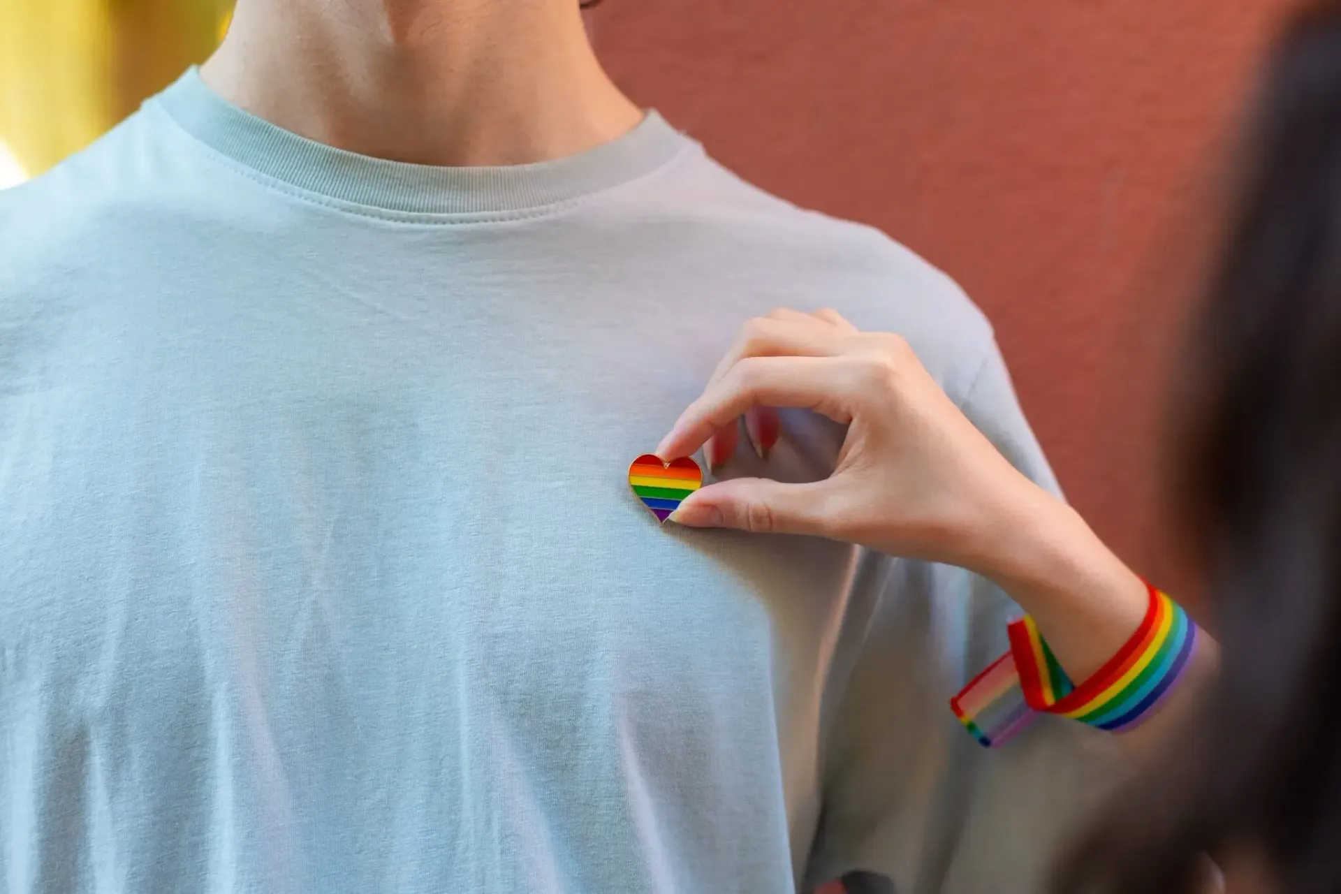 Hand placing a rainbow heart pin on a person's chest, with a rainbow bracelet visible on the right.