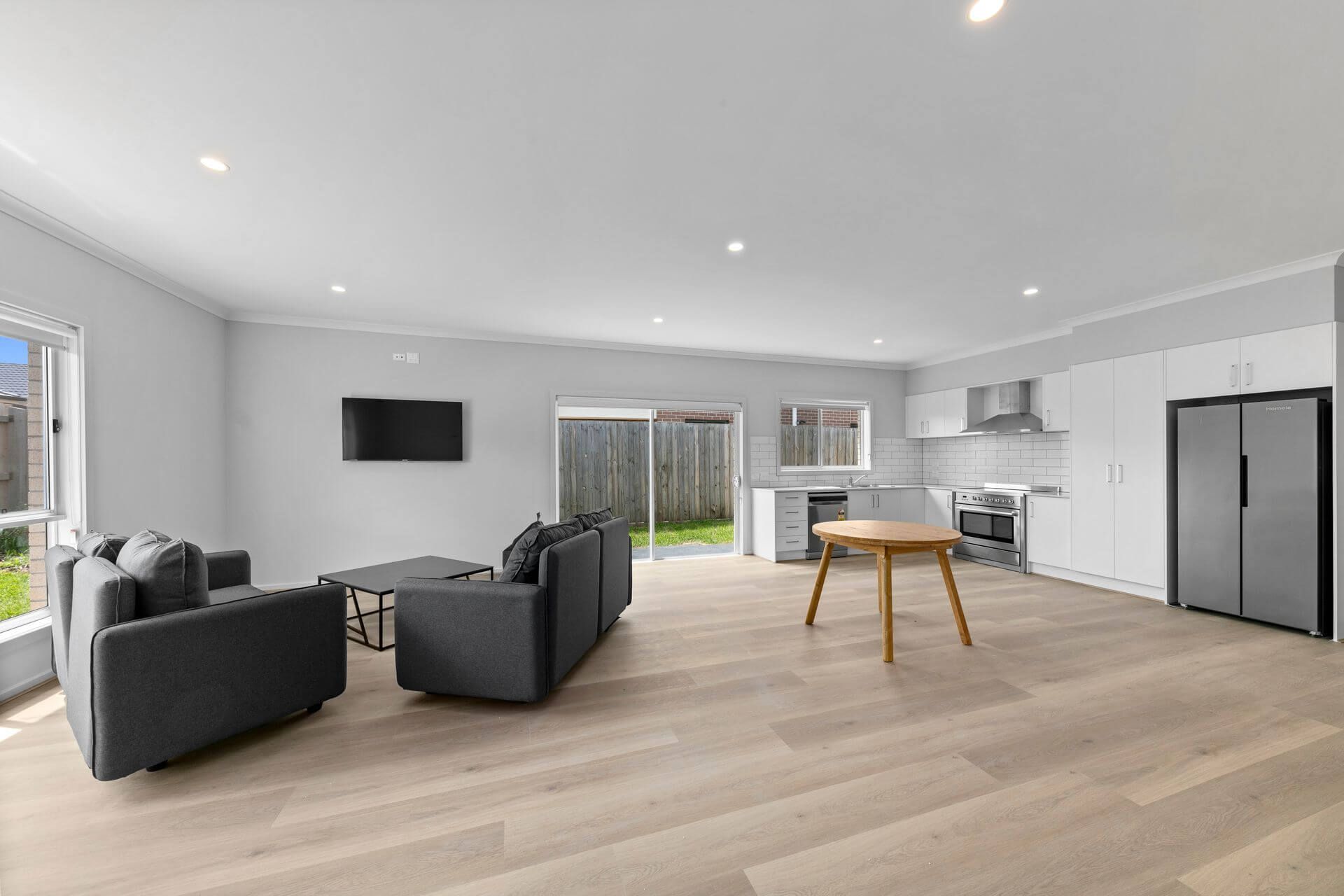 A modern, open-plan living area in Weir Views, VIC 3338 with light wooden floors, two gray sofas, a wall-mounted TV, round wooden table, white kitchen with stainless steel appliances and large refrigerator. Sliding doors lead outside on Marriott Boulevarde.