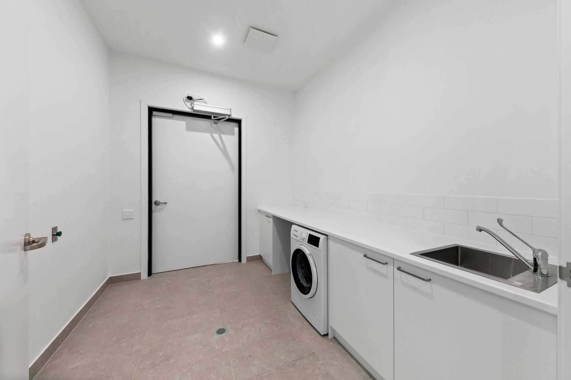 A modern, minimalist laundry room on Serene Terrace, Drysdale VIC 3222, featuring white walls, a washing machine, stainless steel sink, sleek countertop and cabinets. Tiled flooring completes the space with a closed door at the far end.