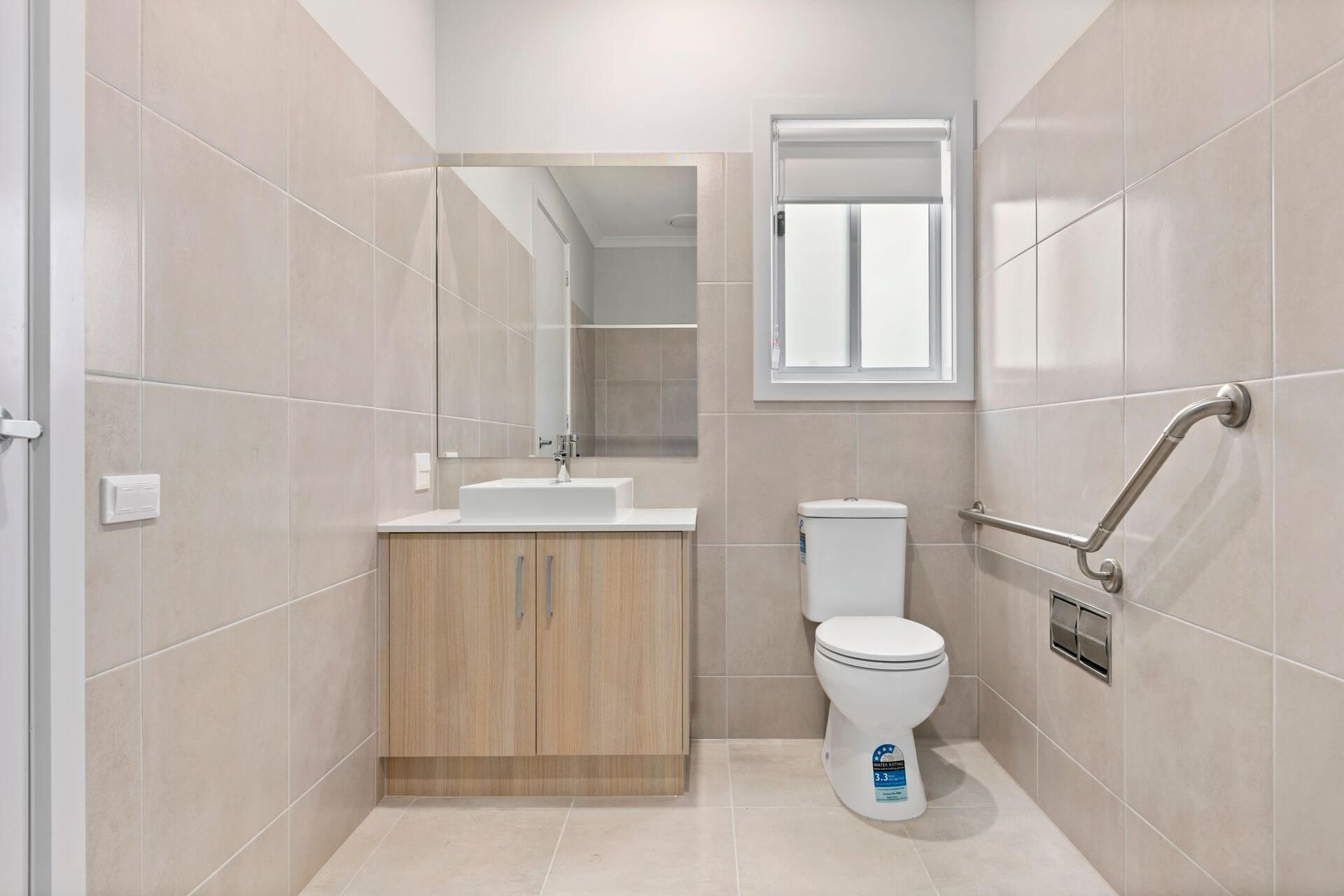 Modern beige-tiled bathroom in the heart of Armstrong Creek, VIC 3217, featuring a wooden vanity and sink, large mirror, white toilet with grab bar beside it, and a frosted window above on Avocet Way.