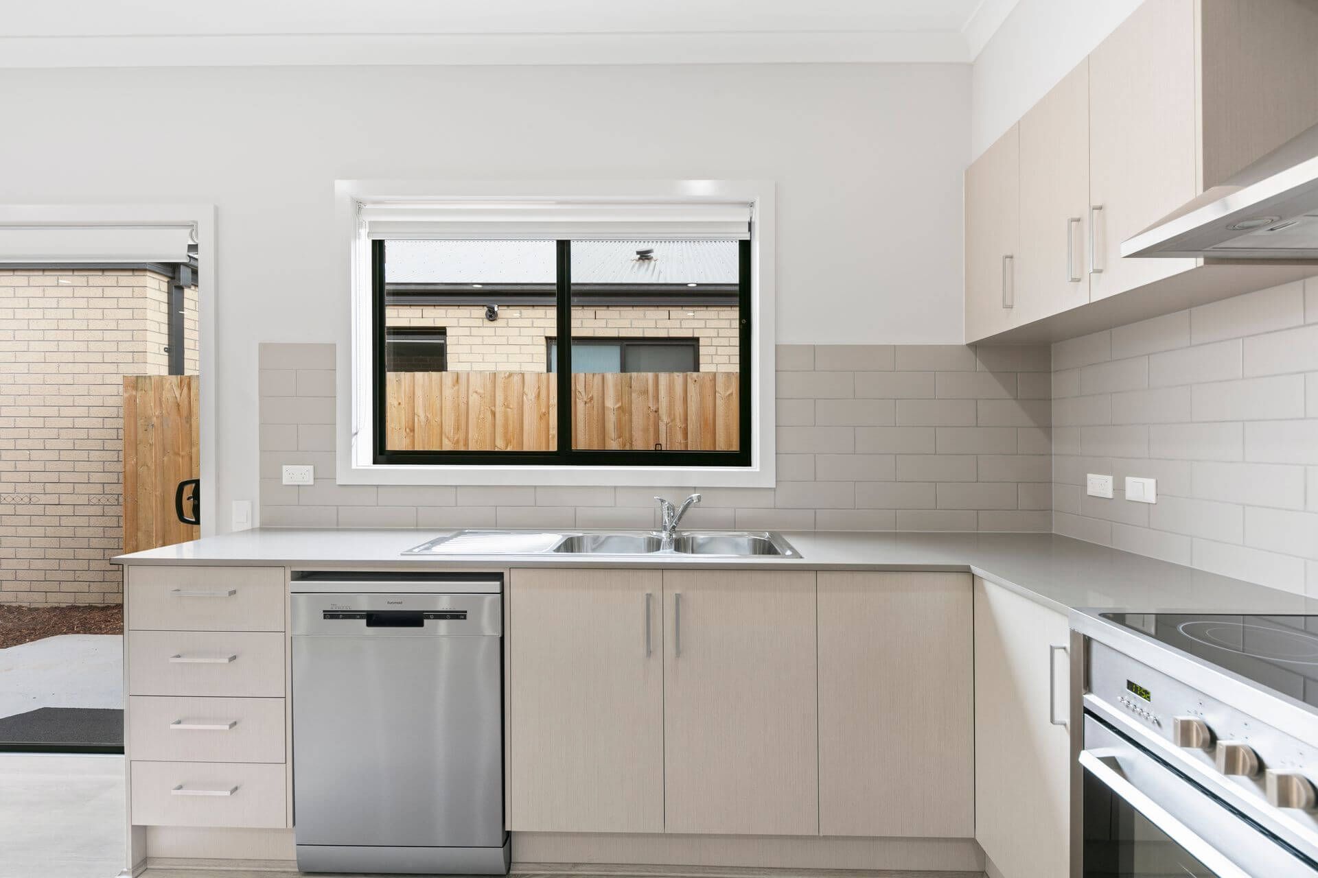 Modern kitchen with light beige cabinets, stainless steel dishwasher, double sink under a window overlooking Marriott Blvd in Weir Views VIC 3338. Features an electric stove and light gray subway tile backsplash; wooden fence and building visible outside.