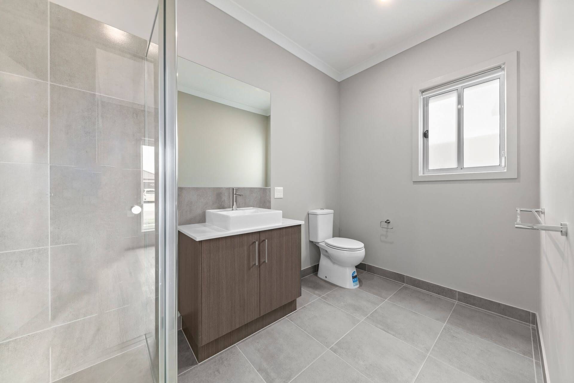 Modern bathroom in Beveridge with gray tile floor, a glass shower on the left, vanity with square sink and mirror in the center, and a toilet next to a window on the right. Walls are painted light gray. Ideal for Magnolia Crescent, VIC 3753 homes.