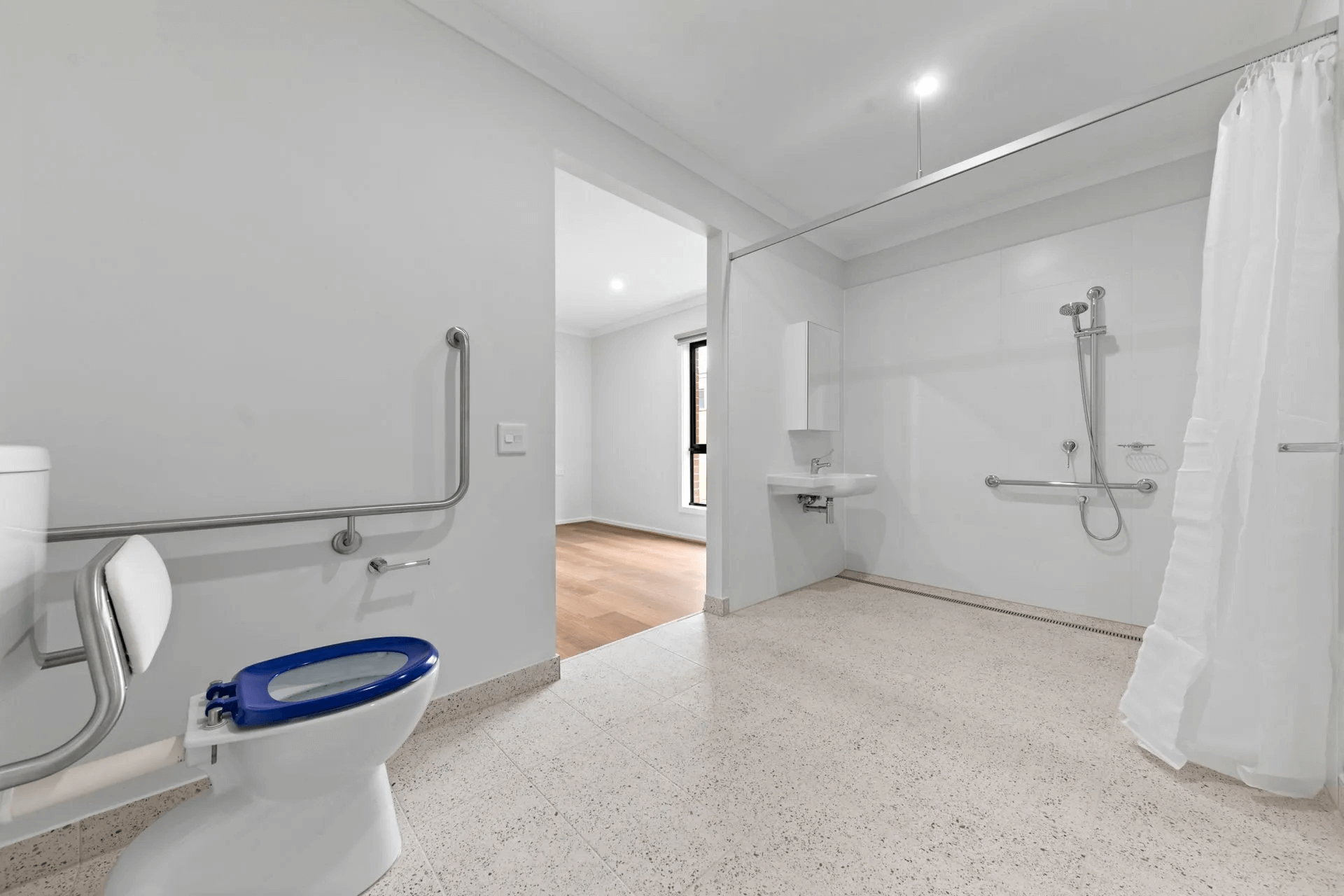 Accessible bathroom on Cygnet Way, Mickleham, VIC 3064 features grab bars, a blue-seated toilet, wall-mounted sink, open shower area with curtain, large mirror, and white walls and flooring. An adjacent space is visible.