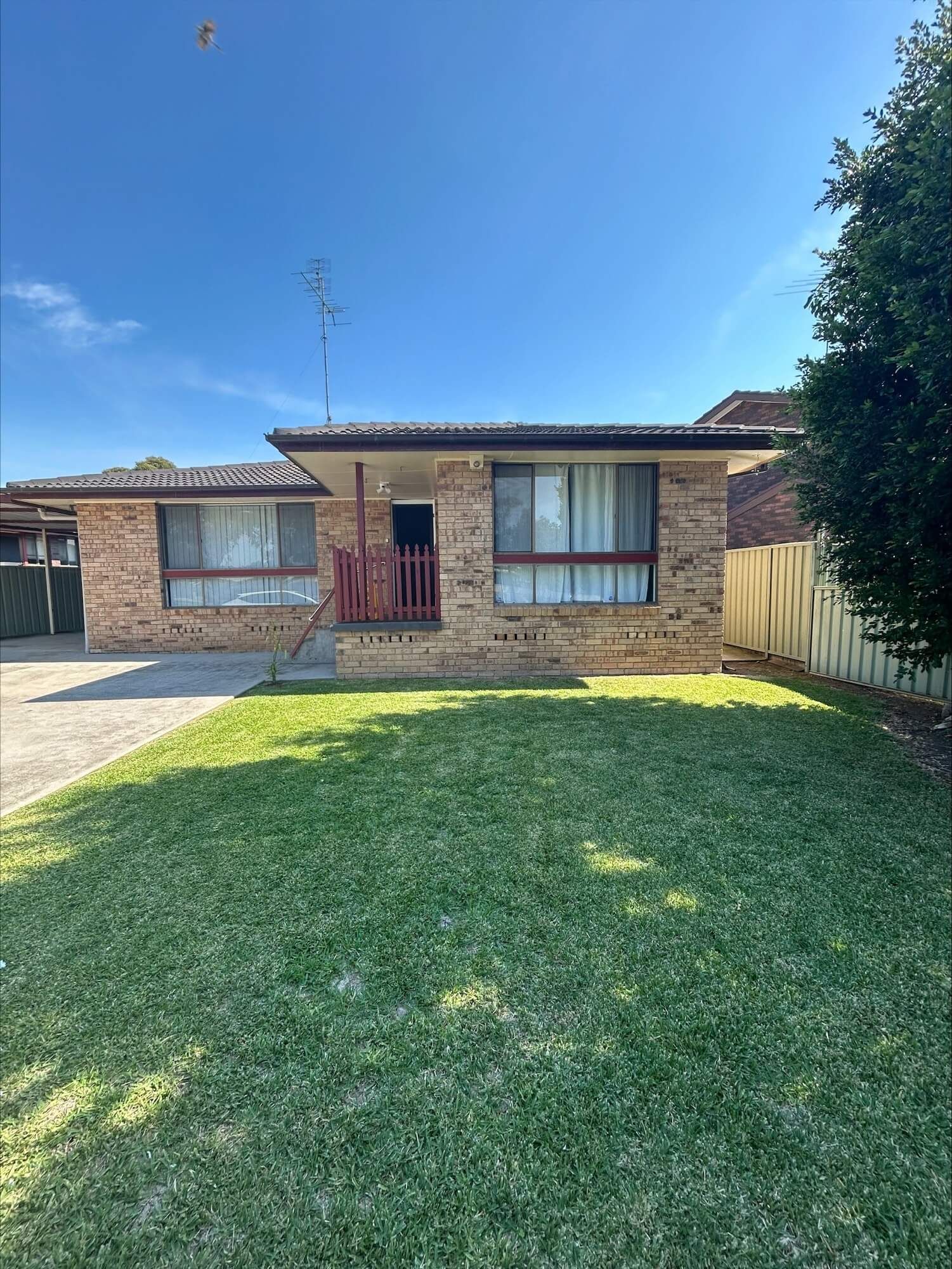 The modern bedroom, part of the Mandalay Estate collection, features a large bed with gray and white bedding on a wooden frame. Two bedside tables with lamps flank the bed. A potted plant and dresser enhance the left side, while a window with blinds and wall-mounted air conditioning complete this elegant space.