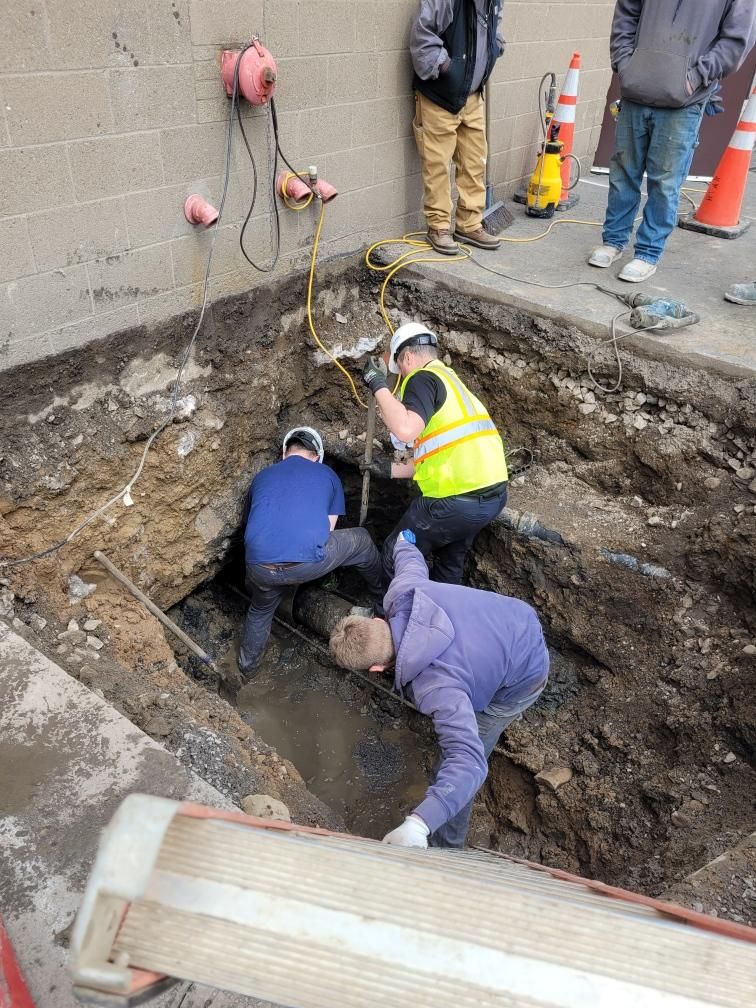 Construction workers in a deep trench, digging and working with tools near a building wall.