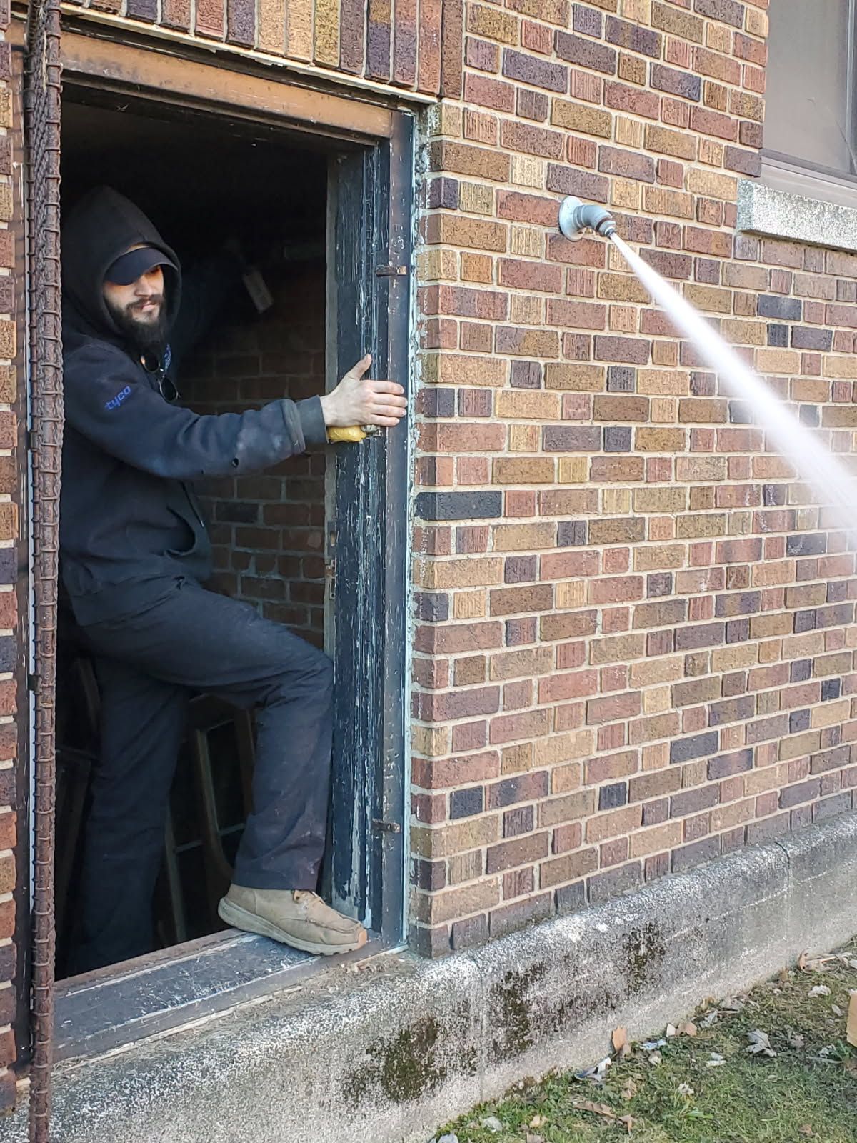 Man in black jumpsuit exits building through doorway, facing a jet of water spraying from a wall-mounted nozzle.