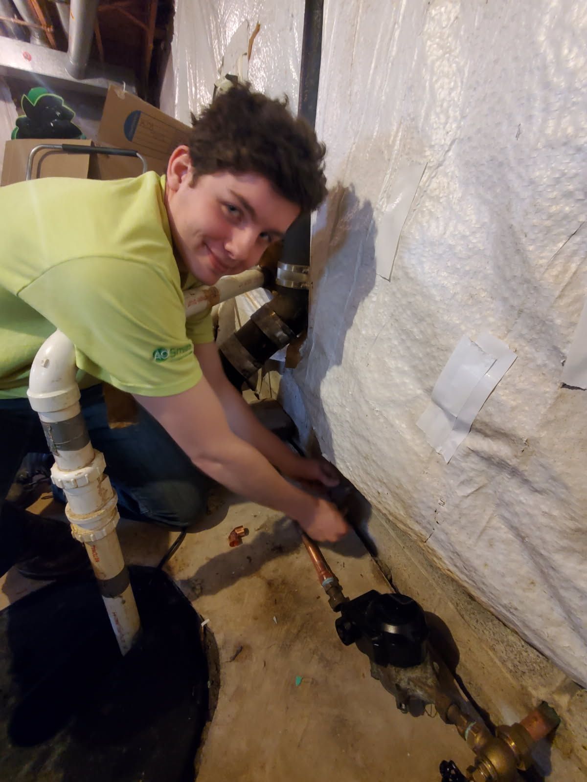Man in green shirt working on plumbing in a basement. Smiling, kneeling near pipes and sump pump.